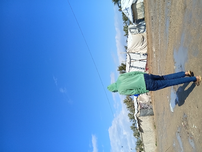 A person in a green hoodie and jeans stands on a muddy ground near several tents and brick structures. The person's back is to the camera, obscuring their face. The ground appears wet with puddles. In the distance, there are palm trees and possibly more brick structures. A vast expanse of blue sky fills most of the frame, with some scattered white clouds towards the right.