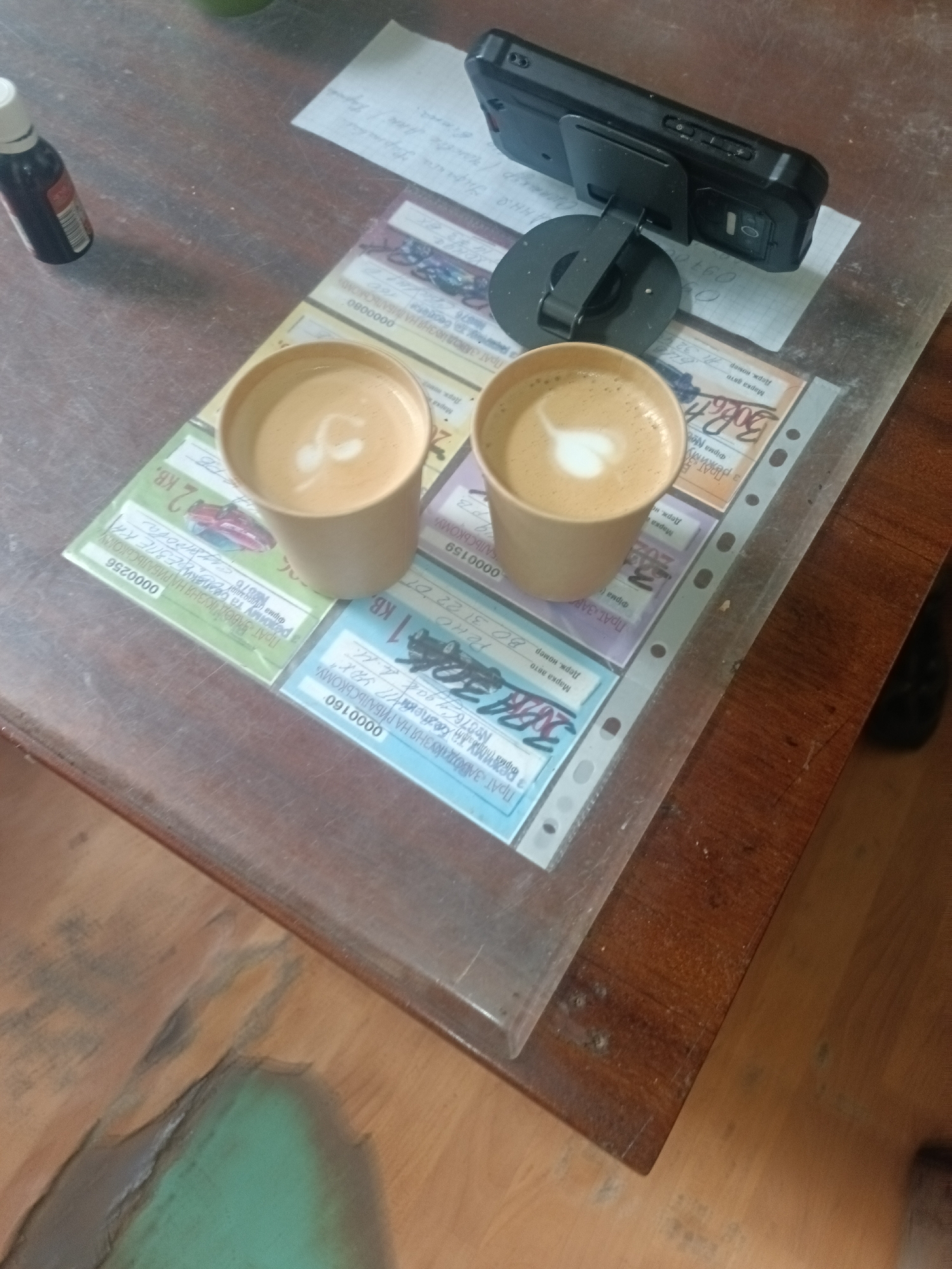 The image depicts a wooden table with two cups of coffee, a smartphone on a stand, and a few papers. The coffee in the cups has latte art. The papers appear to be some sort of documents or tickets with printed text and images. The smartphone is black and is placed on a black stand. The lighting suggests it might be indoors, possibly in a cafe or at home. The overall scene is casual and relaxed. No specific location in Kyiv, Ukraine, can be determined from the image.