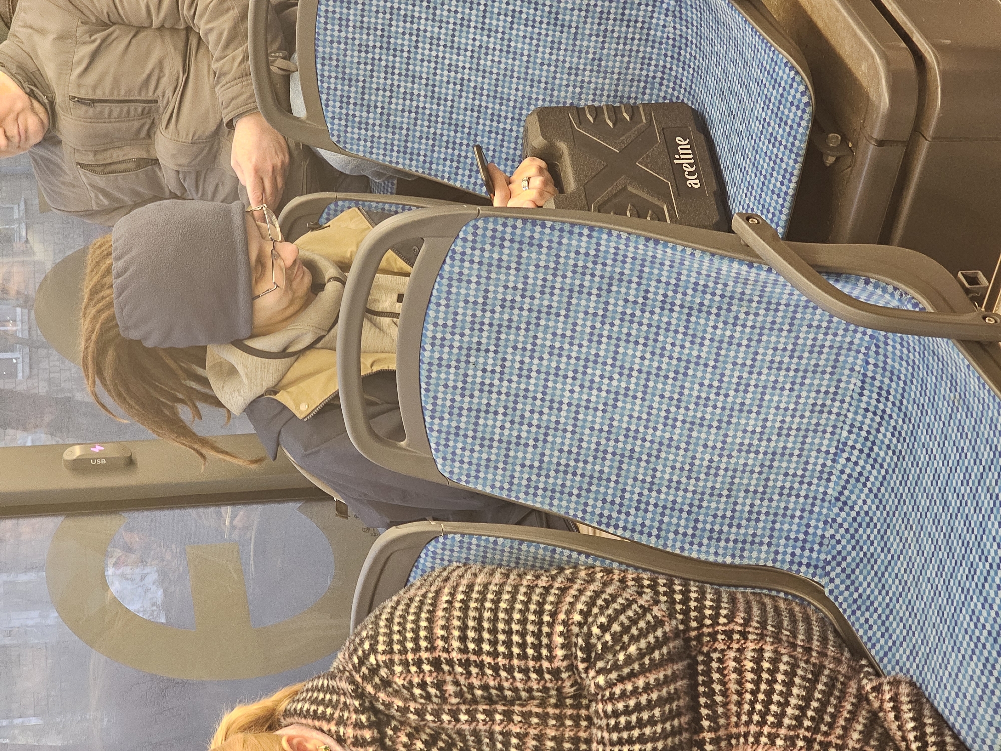 A photograph taken inside what appears to be a passenger transportation vehicle, possibly a train or bus. The vehicle's seats are upholstered with blue and white checkered patterned fabric.
Three passengers are partially visible. One wears a gray fleece hat and has dreadlocks. A second passenger is wearing glasses. A third passenger is wearing a houndstooth-patterned sweater or jacket. Another passenger is in the background on the left side of the photo with their glasses in hand.
A black, rectangular case with the word "aceline" on it is placed on one of the seats. A hand holding a phone is visible near the black case.
A USB port is visible on the side of the vehicle. The image is rotated counter-clockwise. The location is not discernible to Khimki, Russia.