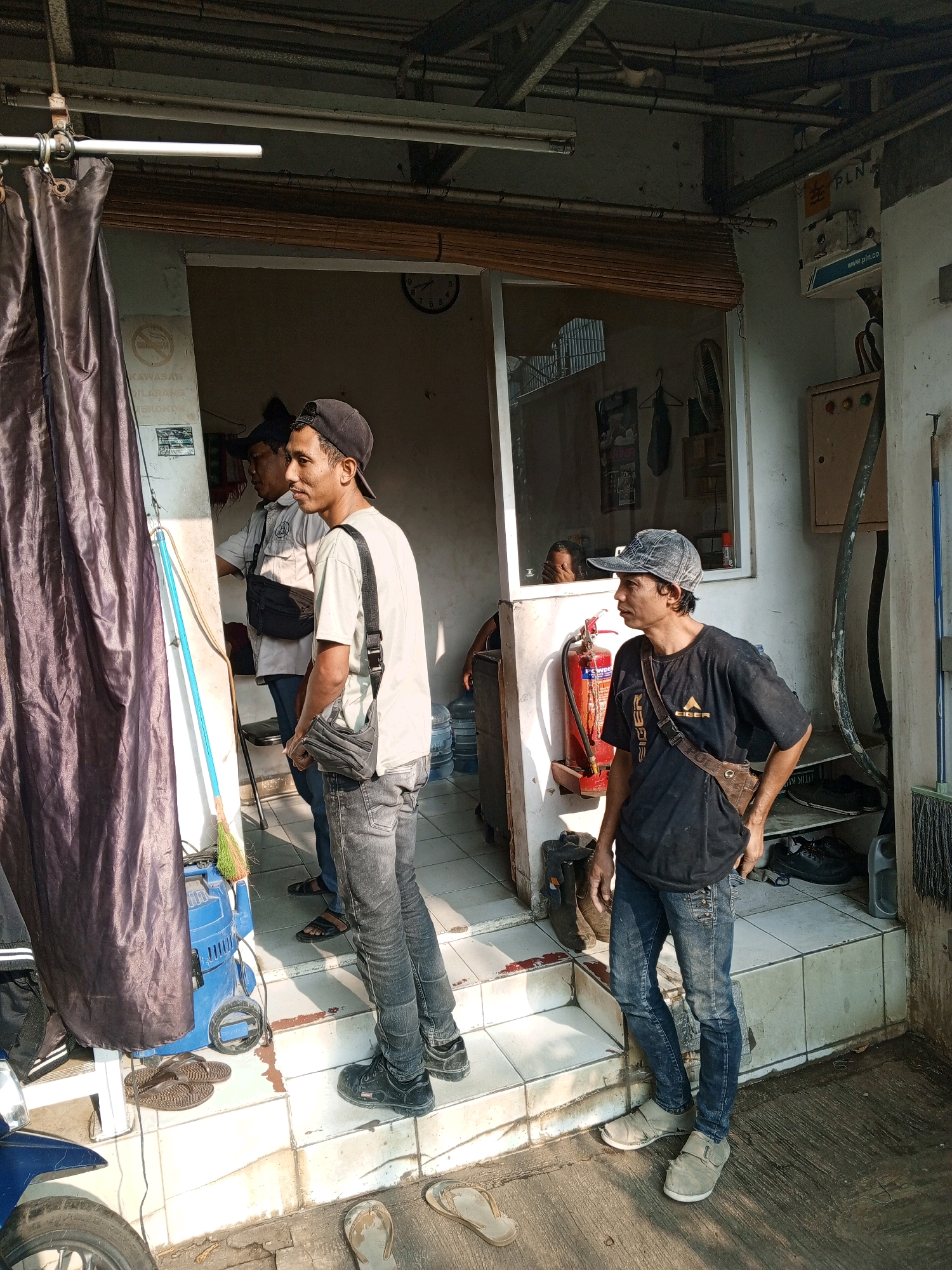 The image depicts an outdoor scene, likely in front of a small shop or service establishment, in the city of North Jakarta, Indonesia. The architecture suggests a commercial area. In the foreground, two men are standing on a tiered entryway. The man on the left is wearing a light-colored t-shirt, dark jeans, and a black cap. He has a black crossbody bag slung over his shoulder. The man on the right is wearing a black t-shirt with a logo, dark jeans, and a denim baseball cap. He also has a brown crossbody bag. Both men appear to be looking towards the right side of the frame.
Behind them, a dark curtain hangs on the left, partially obscuring a blue piece of equipment, possibly a pressure washer, and a broom with green and yellow bristles. Further inside the establishment, visible through a glass window and open doorway, are more details of the interior. There is a fire extinguisher mounted on the wall, a clock on the wall displaying approximately 2:30, and what appears to be a distribution board with the "PLN" logo, indicating the Indonesian state electricity company. Shelving units are visible within the establishment, holding various items, including shoes and possibly containers. The environment appears to be an urban setting, with elements like a concrete floor, tiled steps, and overhead structures. The lighting suggests it is daytime.