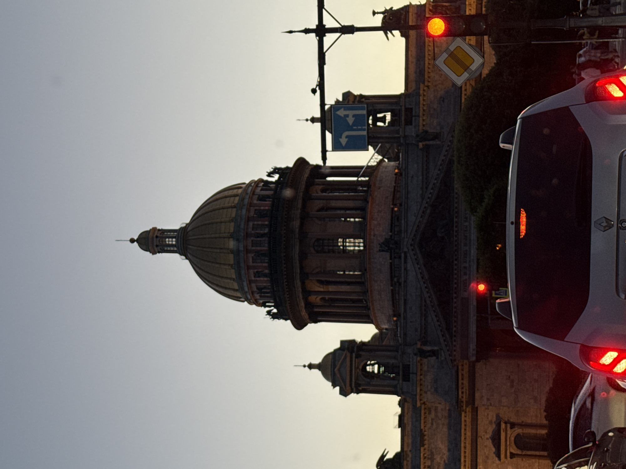 The image depicts Saint Isaac's Cathedral in Saint Petersburg, Russia, at dusk. The cathedral's golden dome and neoclassical architecture dominate the upper portion of the frame, silhouetted against a pale, hazy sky. In the foreground, traffic is stopped at a red traffic light. The rear of a white vehicle, possibly a minivan or SUV, is prominently featured, its taillights illuminated. Other cars are visible in the traffic jam, and pedestrians can be seen walking on the sidewalk in the background. A traffic light with a red illuminated bulb and a diamond-shaped road sign are also visible. The scene suggests a typical urban setting with heavy traffic congestion during the evening hours.