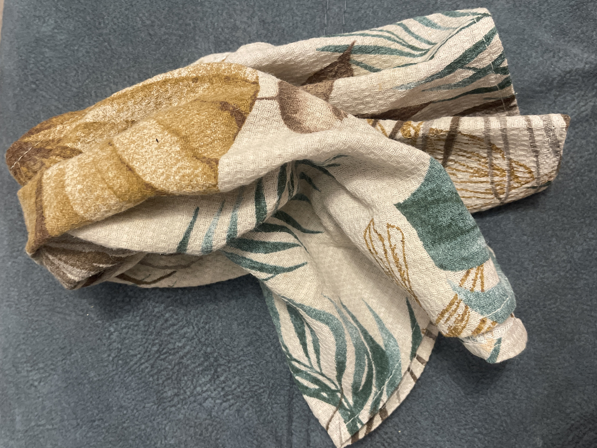 A crumpled, light-colored towel with a pattern of green leaves and brown and tan abstract shapes is resting on a dark blue, textured surface. The towel is folded and twisted, with its edges visible. The pattern on the towel is abstract and organic, with flowing lines and soft colors. The dark blue surface appears to be fabric, possibly a couch or chair. The lighting is natural and casts soft shadows.