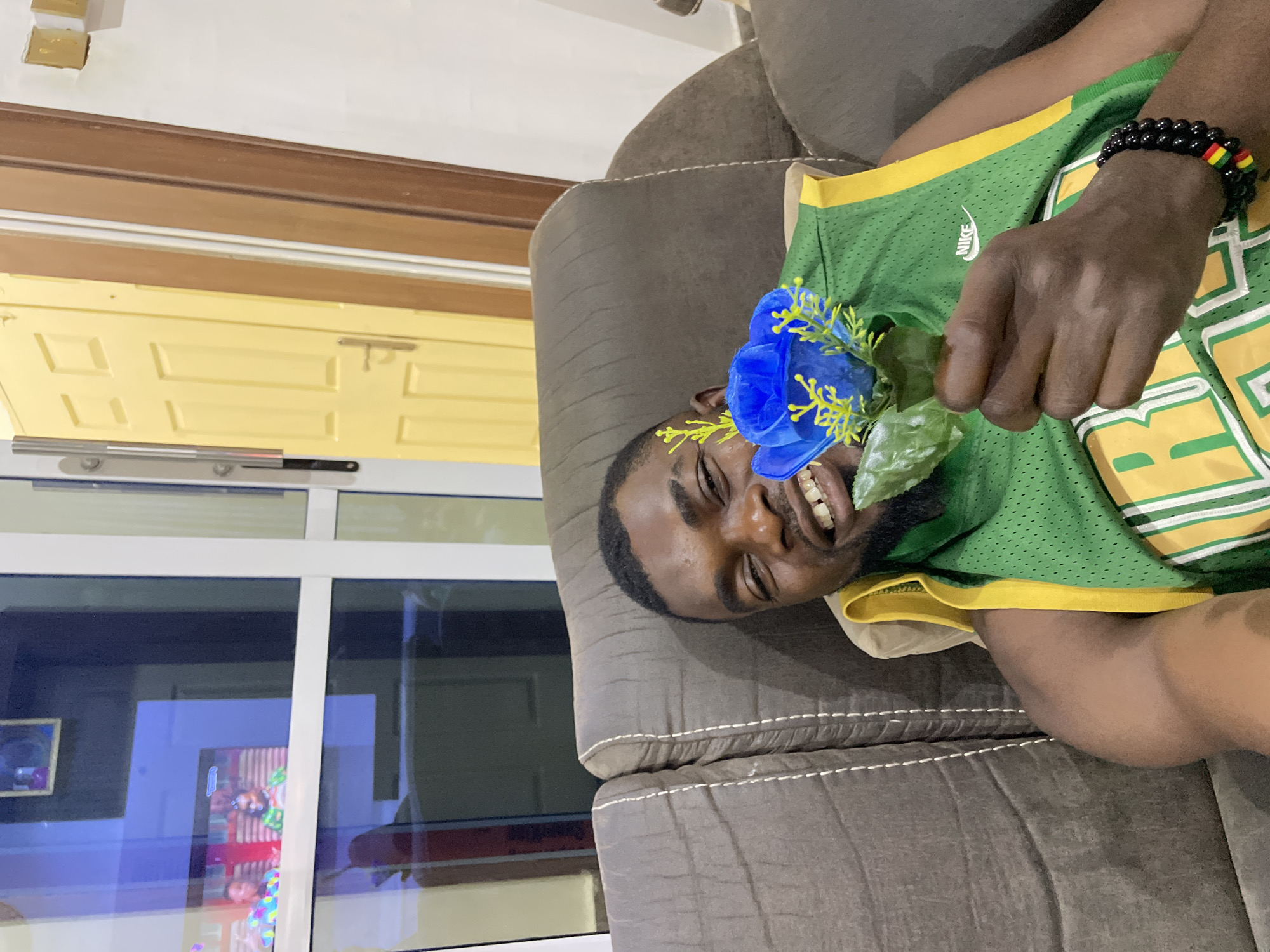 A medium, full shot depicts a dark-skinned man lying on a brown couch. He is wearing a green and yellow basketball jersey. He holds a blue rose with green leaves in his hand. He is smiling, and a bracelet is visible on his wrist. In the background, a yellow door is partially visible, as well as a glass window reflecting images of an interior room. The scene likely takes place indoors. The location is specified as Jos, Nigeria.