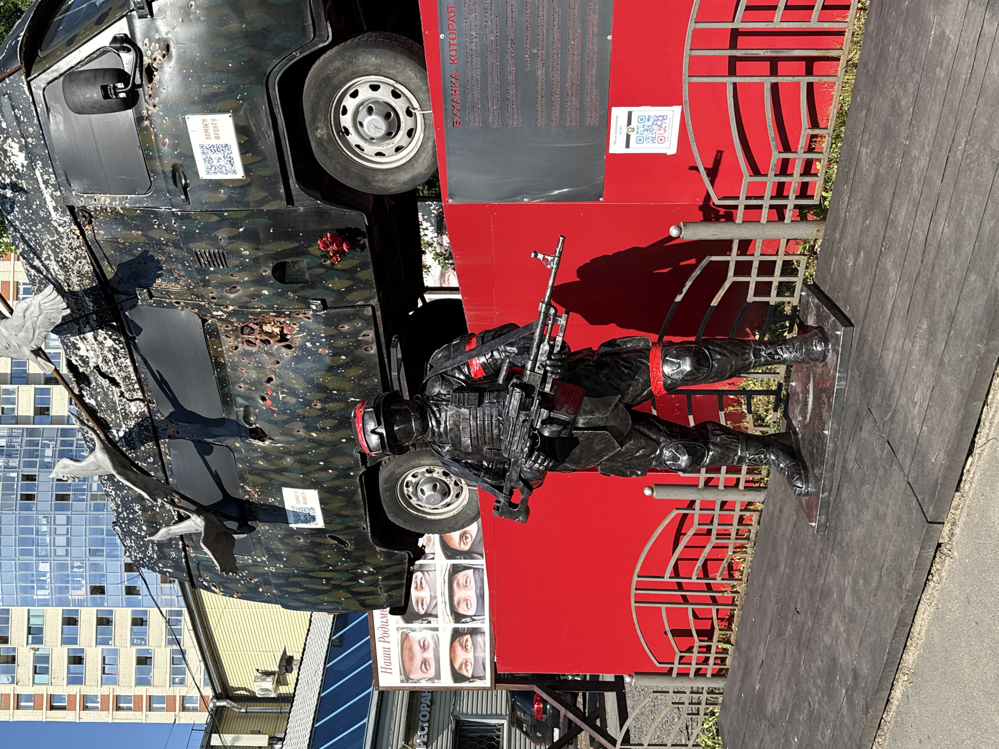 This media file depicts an outdoor display featuring a damaged vehicle and a statue of a soldier. The vehicle, a dark-colored van with camouflage paint and numerous bullet holes, is positioned at an angle, as if it has been partially flipped or is part of a larger exhibit. A QR code and a sign with Cyrillic text are affixed to the side of the van.
In front of the vehicle, a life-sized statue of a soldier in black tactical gear holds a rifle. The soldier is standing, poised, with a metallic sheen to its armor. The display is set against a vibrant red background, bordered by a decorative metal fence.
To the left of the display, a modern glass building stands, suggesting an urban environment. Below the van, a series of portraits of people's eyes are displayed on a board, accompanied by Cyrillic text. A sign for a restaurant is also visible. The lighting suggests it is daytime, with clear weather. The overall scene appears to be an artistic installation or memorial, possibly related to a military event, given the damaged vehicle and the soldier statue. The text on the display elements mentions "Front," "Our Homeland," and "Bukhanka," which is a nickname for the UAZ-452 van.