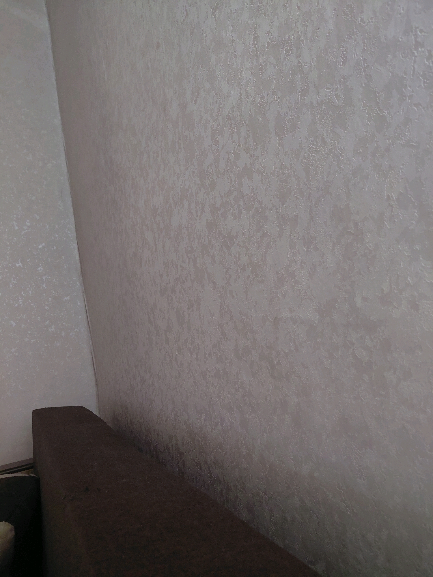 The image shows an interior corner of a room. A dark brown upholstered object, possibly a sofa or a bed, occupies the lower left portion of the frame. The walls are covered in a textured, light-colored wallpaper. The wallpaper has a subtle, abstract pattern. The lighting suggests an indoor setting, with the light source likely coming from the right. No people or specific identifying landmarks are visible in the image. The location in Malinovka, Russia, is not discernible from the visual information provided.
