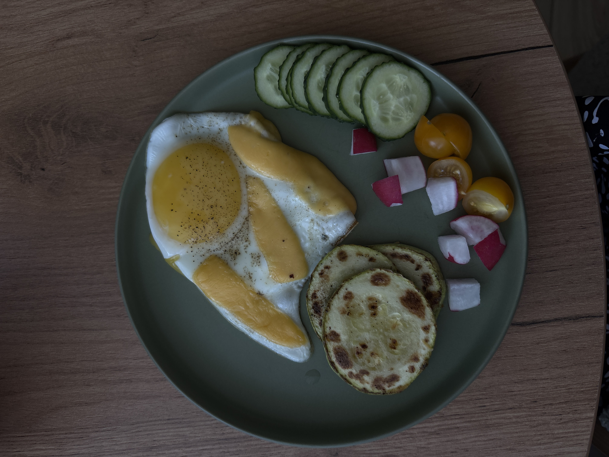 A flat lay shot captures a prepared meal arranged on a round, light green plate, resting on a light brown wooden surface. The meal consists of various components. At the center, there are fried eggs; one prominently displays a yellow yolk and white, generously seasoned with black pepper. Another egg, or part of the same one, features melted, elongated strips of yellow cheese on its white.
Arranged across the top of the plate are several overlapping slices of fresh cucumber. To the right of the eggs, small, irregular cubes of red and white radish are scattered, alongside several yellow cherry tomatoes, some whole and some halved. Below the eggs and radishes, three round slices of what appear to be pan-fried zucchini or squash are visible, exhibiting some browned areas from cooking.
The wooden surface beneath the plate displays a subtle grain pattern. In the very far right edge of the frame, a dark fabric with a white, abstract pattern can be partially seen, possibly indicating a chair or piece of clothing. The lighting suggests an indoor setting, and the meal appears ready for consumption. There is no visible text in the image.