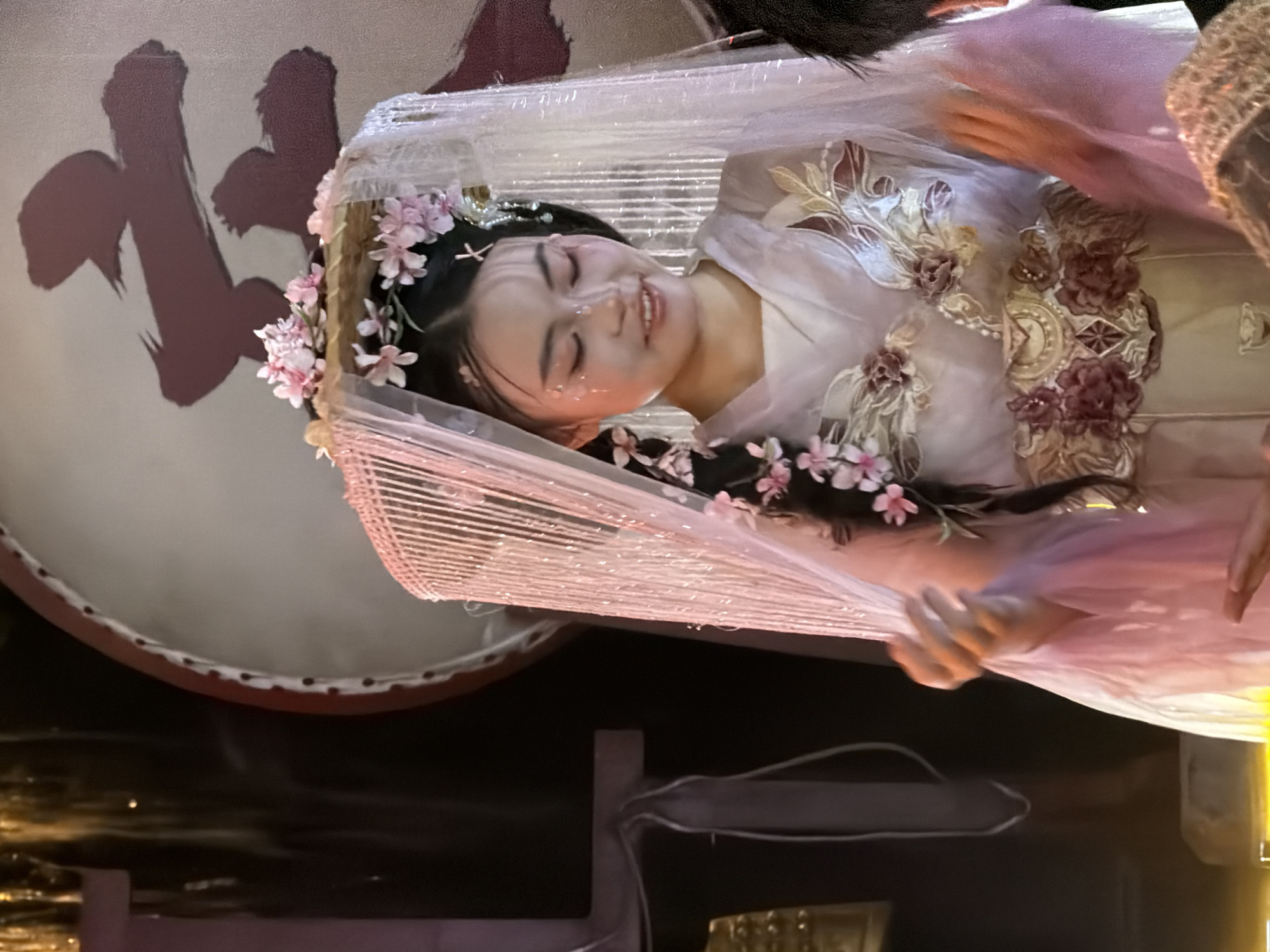 In a dimly lit indoor setting, possibly a performance venue in Yuzhong District, China, a young woman with her hair styled in a braid adorned with pink flowers is captured in a medium shot. She wears an elaborate costume featuring a light purple, sheer outer layer with intricate floral and leafy embroidery in shades of pink, burgundy, and gold. A delicate, sheer veil with hanging strings and scattered pink flowers rests on her head, partially obscuring her face. Her makeup includes a soft pink blush and shimmering accents on her cheeks and forehead. Her hands are positioned near her chest, with her right hand gesturing outwards. In the background, a large, circular drum with a red rim is visible, bearing a dark, calligraphic symbol. To the left, a dark structure with decorative elements and hanging tassels can be seen, along with what appears to be a bell. The overall ambiance suggests a theatrical or ceremonial performance.