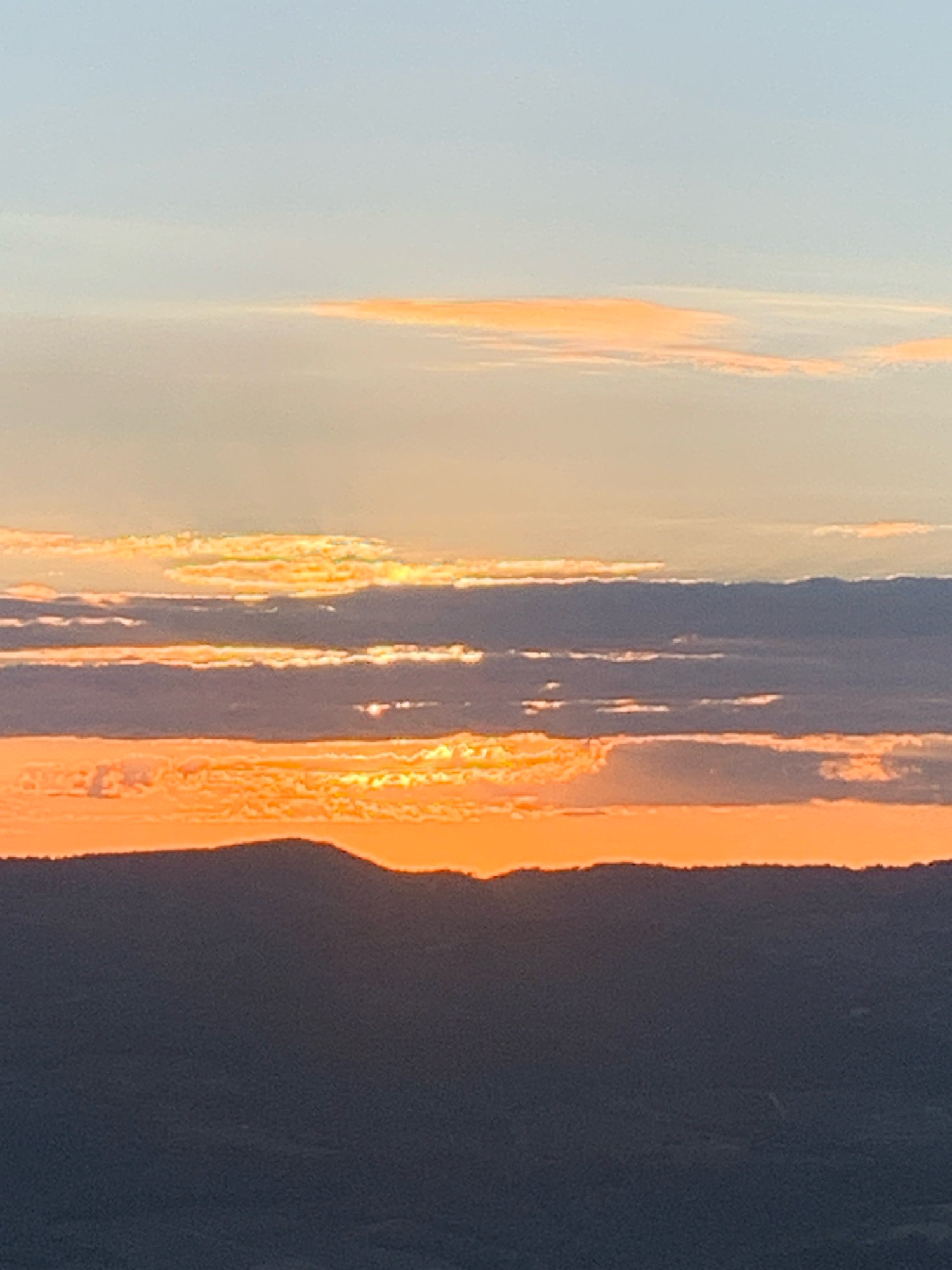 This is a photograph of a sunset over a silhouetted landscape. The sky is a gradient of pale blue at the top, transitioning to soft oranges and yellows as it meets a band of clouds. Below this, there are layers of darker, wispy clouds, some illuminated by the setting sun with bright orange and yellow edges. The horizon is marked by the dark silhouette of rolling hills, with a distinct peak in the center. The foreground is a darker, indistinct shadow of the landscape. The image appears to be taken from a high vantage point, looking out over a valley or expanse. The lighting suggests the golden hour, with the sun low on the horizon. The location is Castiglione d'Orcia, Italy.