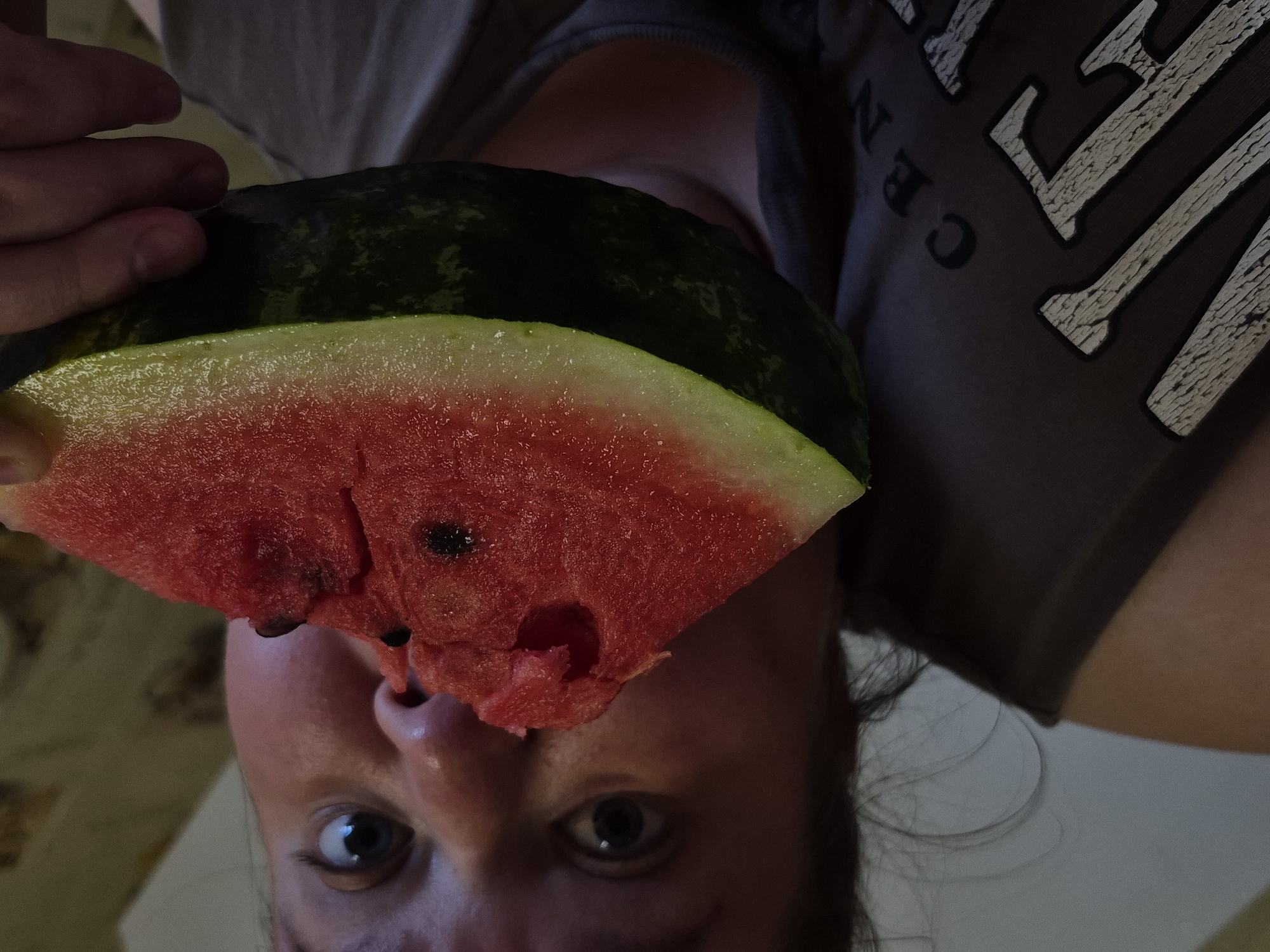 A person is holding a slice of watermelon to their face, obscuring much of their features. The person's eyes are wide open, and their mouth is partially visible, appearing to be in the process of eating or about to eat the watermelon. The watermelon is a vibrant red on the inside with visible seeds, and a green rind. The person is wearing a dark t-shirt with some text on it, which is partially obscured by the watermelon. The background appears to be an indoor setting with neutral-colored walls. The scene is shot from a low angle, looking up at the person and the watermelon. There is no discernible information about the location within the city of Дойная, Russia.