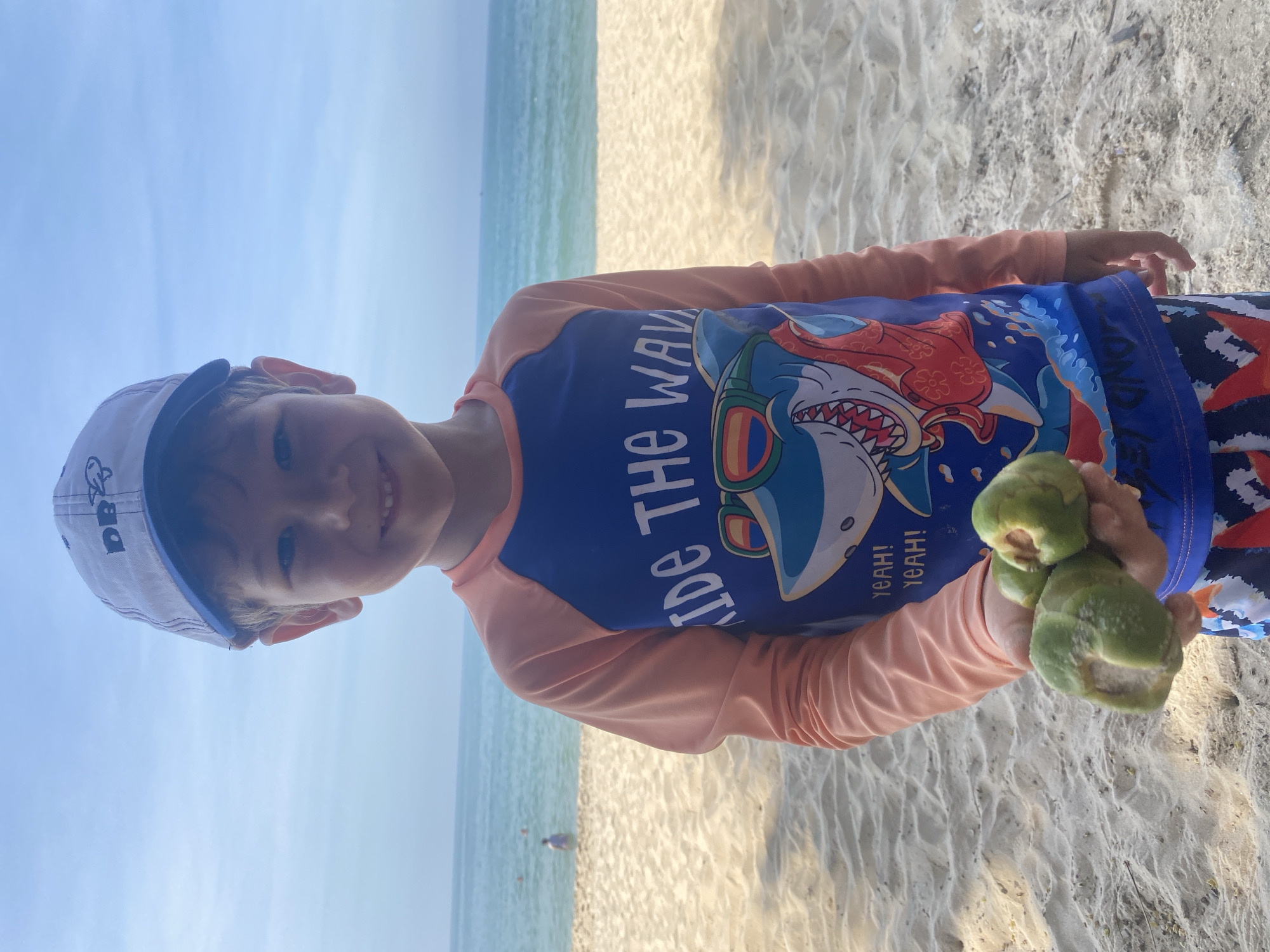 A young boy wearing a white cap with blue trim and a blue and peach rash guard with a shark graphic stands on a sandy beach. He is holding three small, green coconuts in his right hand. The ocean is visible behind him, with a few people in the water and a gentle surf breaking on the shore. The sand is light-colored and appears dry. The sky is a clear, pale blue. The boy is smiling and looking directly at the camera. The scene appears to be in Phu Thuy Ward, Vietnam.