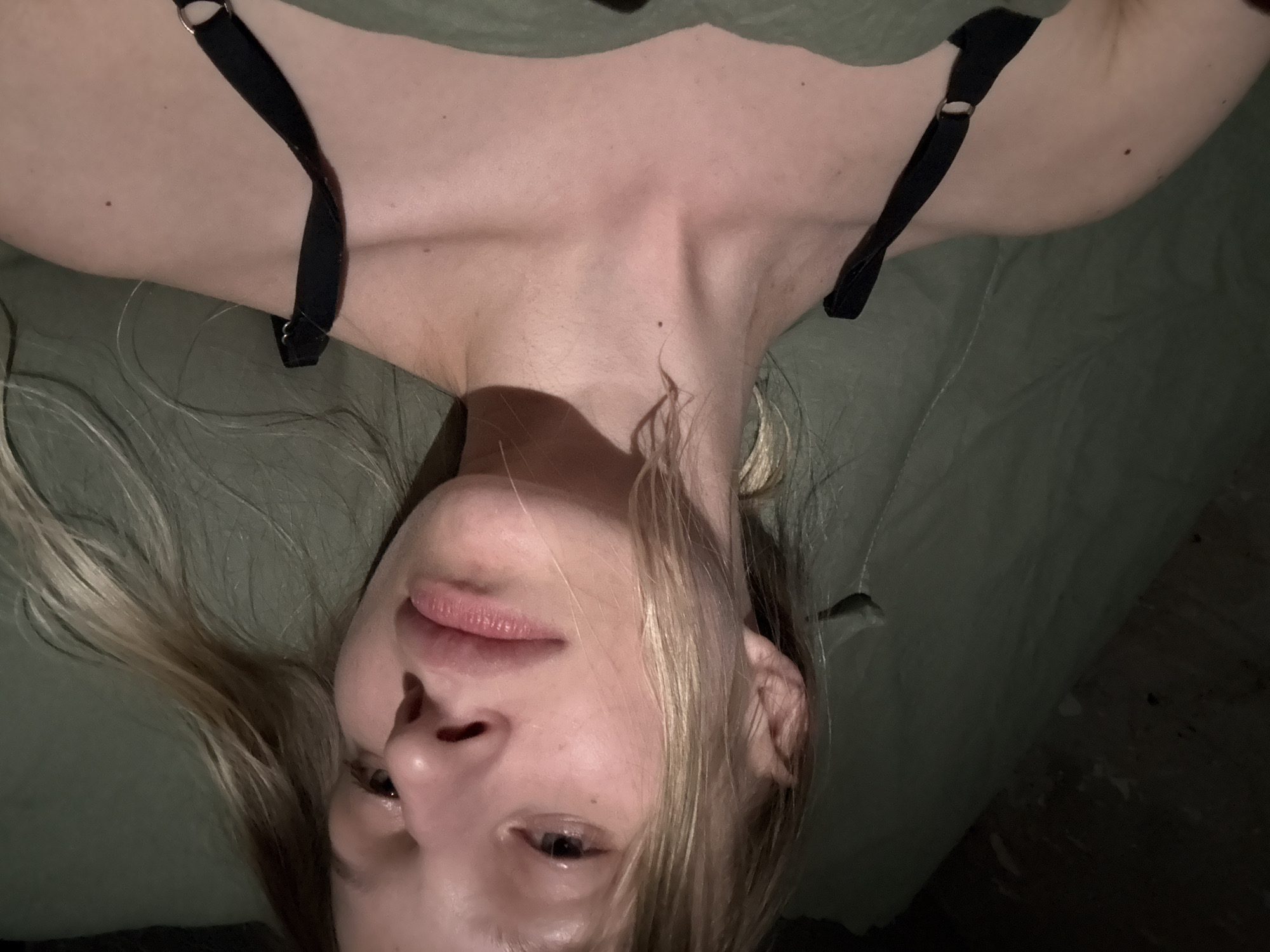 An overhead shot depicts a blonde woman lying on a green surface. She is wearing a black garment with straps. The woman's face is partially illuminated, revealing her nose, lips, one eye, and part of her hair. The background is dark and unclear. There are no details to determine the location of the image, nor its relation to the city of Volkovka, Russia.