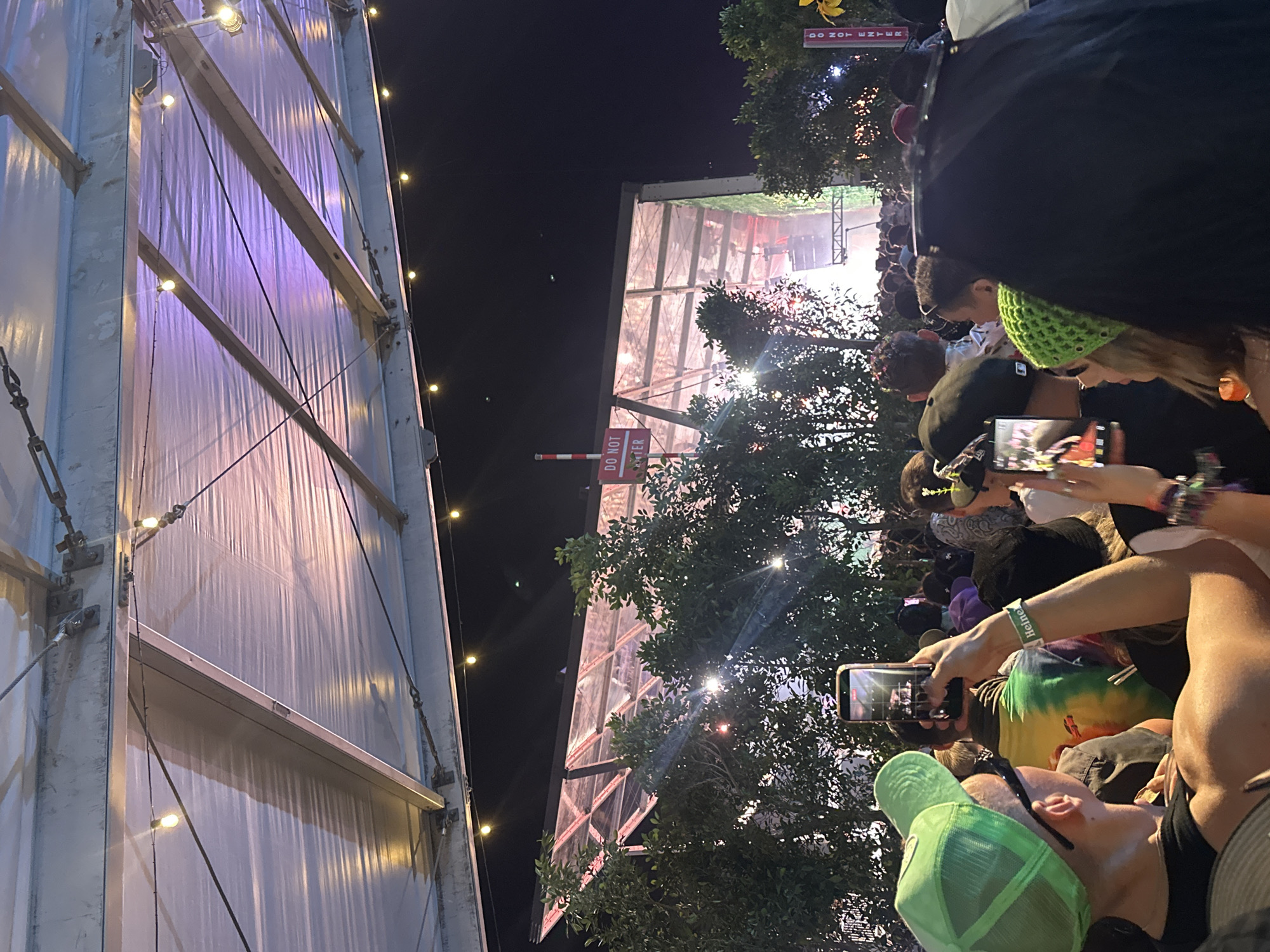 Captured at an outdoor music event in Unknown, Unknown, the image reveals a dense crowd of concert attendees. The composition is vertical. The foreground is dominated by concert-goers, many holding up their smartphones to record the performance, as is evidenced by the screens displaying the stage. Some people are wearing baseball caps and there is a green crocheted beanie and a neon green baseball cap. In the background, a structure, illuminated by what appears to be stage lighting, stands tall behind the crowd and is partially obstructed by tree branches. A sign reading "DO NOT ENTER" is also visible, placed amidst the tree cover. The overall atmosphere suggests a lively music venue at night, teeming with participants.