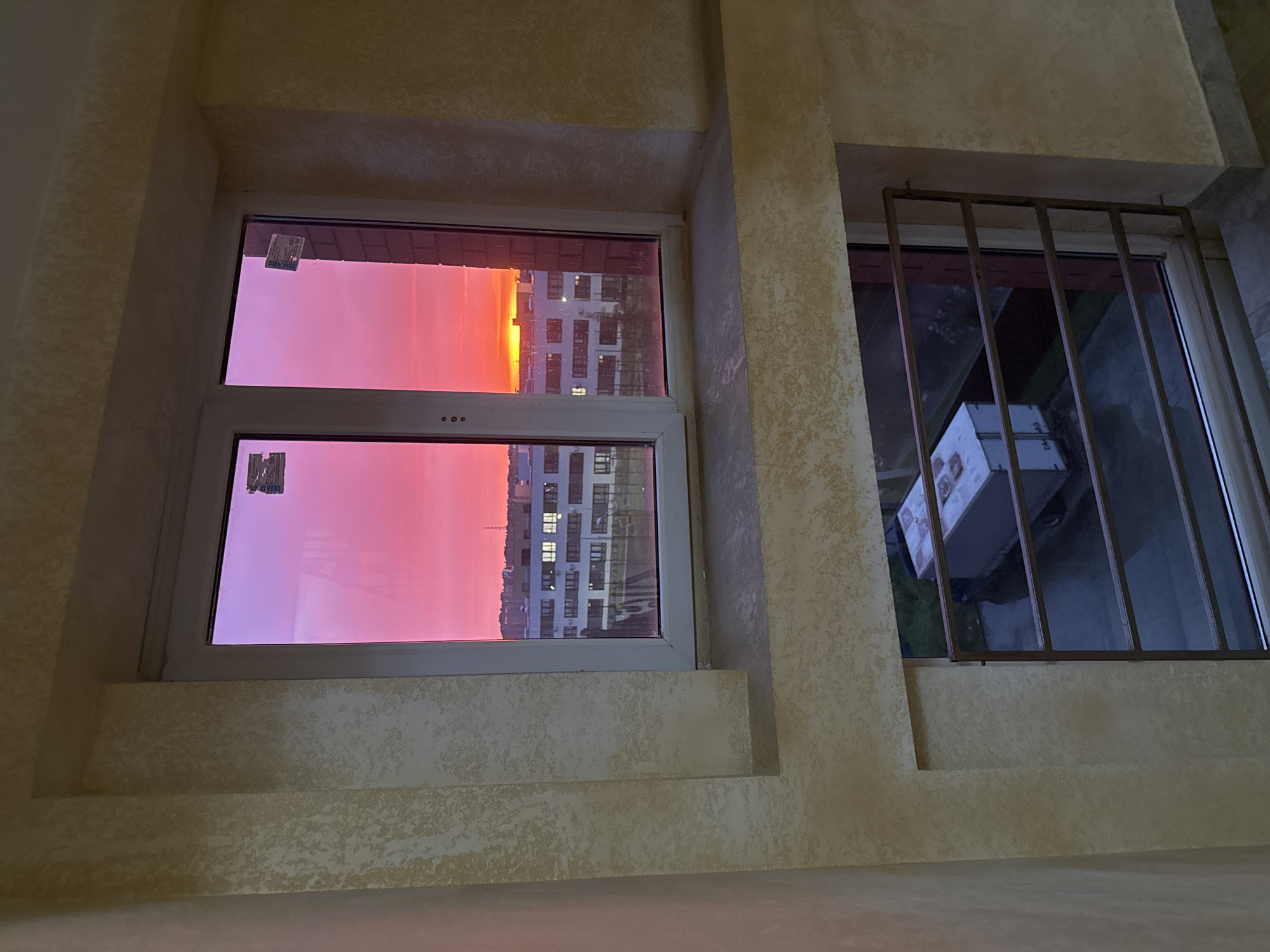 The image shows two windows within an interior wall, likely within a building in the city of Unknown, Unknown. The window on the left reveals a cityscape bathed in a vivid pink and orange sunset. A tall building is visible with multiple illuminated windows. The window on the right has a metal security grille. Through the grille, there's a partial view of a white cardboard box. The wall surrounding the windows has a textured, patterned finish.