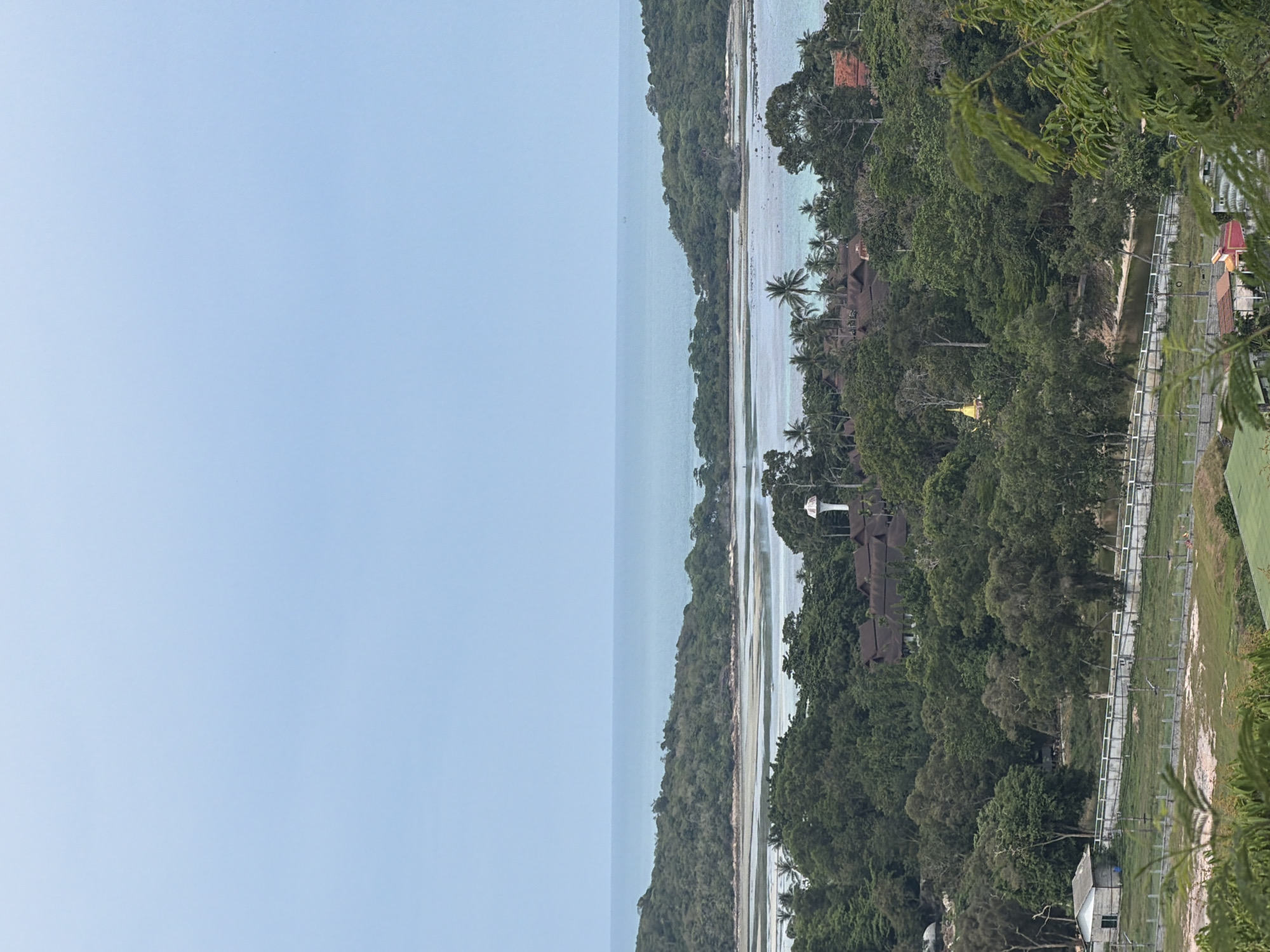 The image captures a panoramic view of a coastal landscape, likely in Baan Bang Rak, Thailand. The scene is dominated by a serene blue sky meeting the tranquil turquoise ocean. In the mid-ground, a lush green island, densely covered with trees, juts out into the water, creating a natural bay. The shorelines are a mix of sandy beaches and rocky outcrops.
Closer to the foreground, a verdant forest blankets the rolling hills. Scattered amongst the trees are glimpses of human habitation. Several buildings with dark, pitched roofs are visible, suggesting a village or resort. A prominent white water tower stands tall amidst the foliage. To the left, a small, light-colored building with a simple rectangular structure is situated near a white concrete wall and fence, with a grassy field beyond. A pathway or road with a series of lampposts runs along the fence line. In the far distance, the ocean stretches towards the horizon. The overall impression is one of a peaceful, tropical setting, with a blend of natural beauty and human presence.