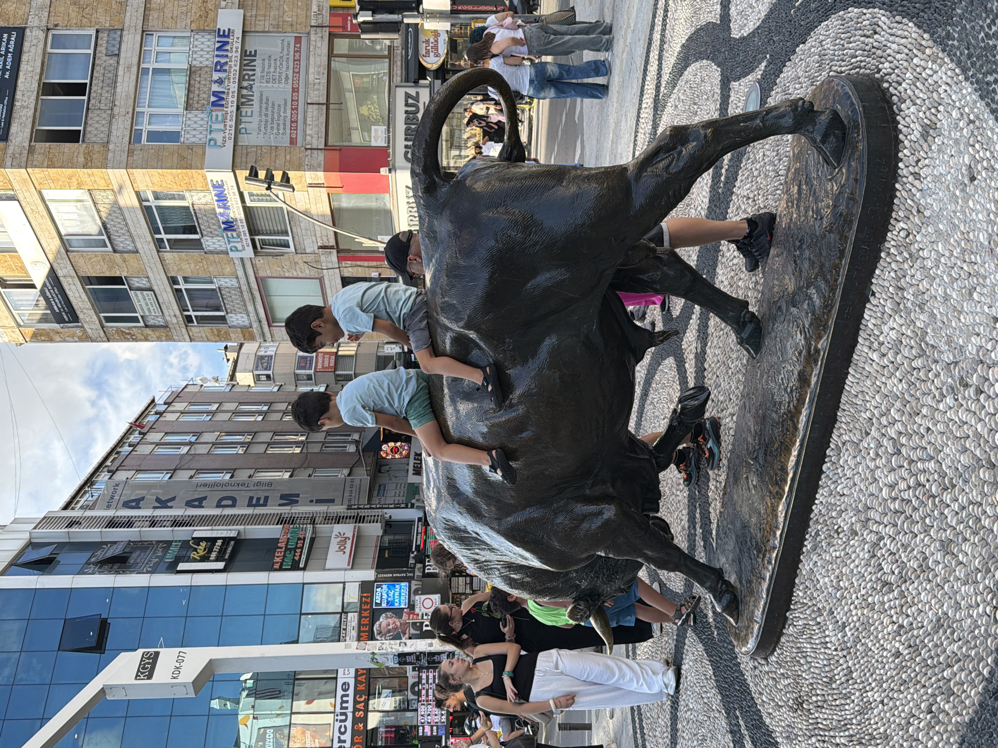 In Kadıköy, Turkey, a bronze bull statue is the central focus of this street scene. Two young boys are seated on the bull's back, one on its hindquarters and the other closer to its head. Several adults are present around the statue, some observing, one standing behind the bull, and others walking in the background. The statue is situated on a ground decorated with small pebbles arranged in a circular pattern. In the background, there are multi-story buildings with various signage, including "KGYS," "AKADEMİ," and "PTEMARINE." A traffic light is also visible on the right side of the image. The sky above is partly cloudy.