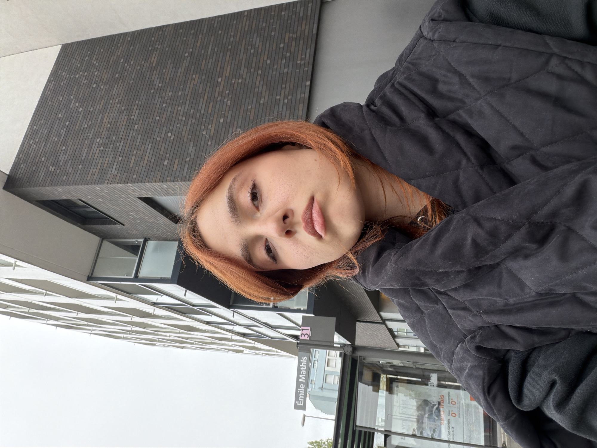 A young woman with auburn hair is facing the camera. She is wearing a black quilted jacket. Behind her is a building with a grey brick facade. In the foreground is a street sign that reads "Émile Mathis 31". The location is in Strasbourg, France.