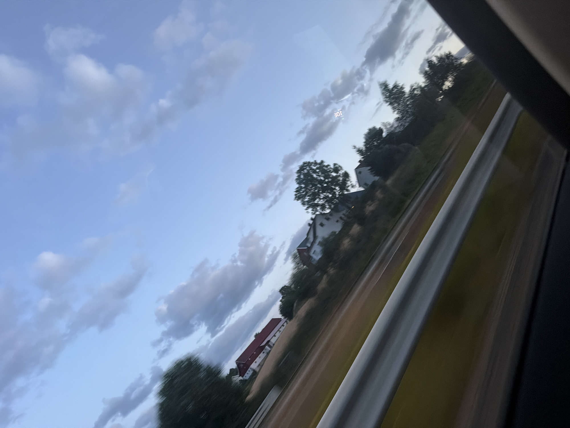 The image depicts a view from a moving vehicle, likely a car, on a road. The scene is characterized by motion blur, indicating a high speed of travel. In the background, there is a rural landscape with several houses and trees. The sky is partly cloudy, with a mix of blue sky and grey clouds. The overall impression is of a scenic drive through the countryside. There are no people visible in the image. The location is not specified as Sarpsborg, Norway.