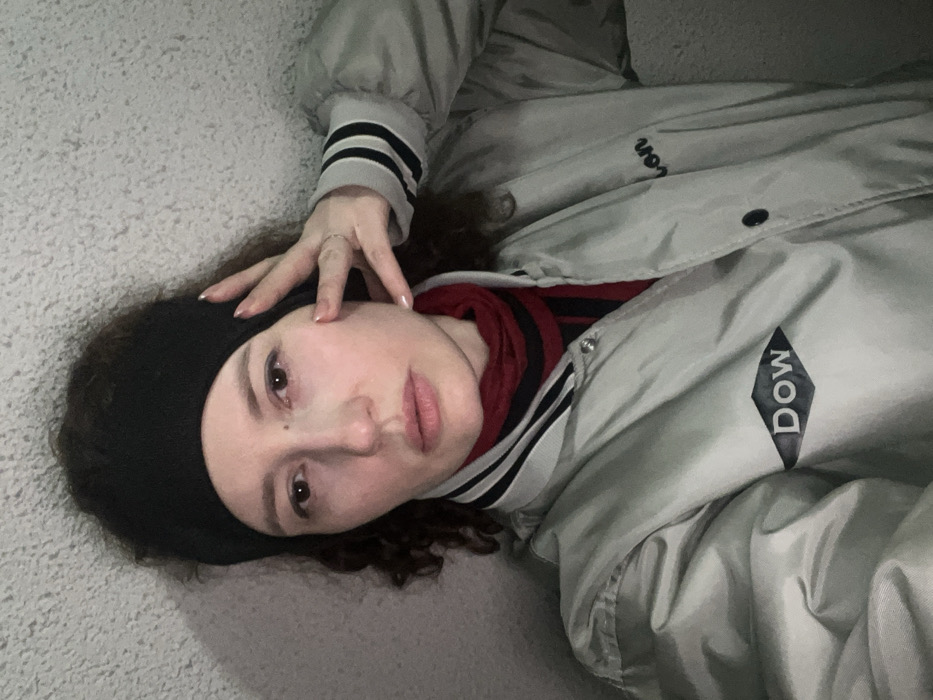 A person lies on a textured, light-colored surface, wearing a black headband and a grey jacket with black and white striped cuffs. The jacket has the word "Dow" on the upper right side, next to a diamond shape. A red and black striped shirt is visible beneath the jacket. The person's left hand rests on their head near the headband. The subject is gazing towards the camera.