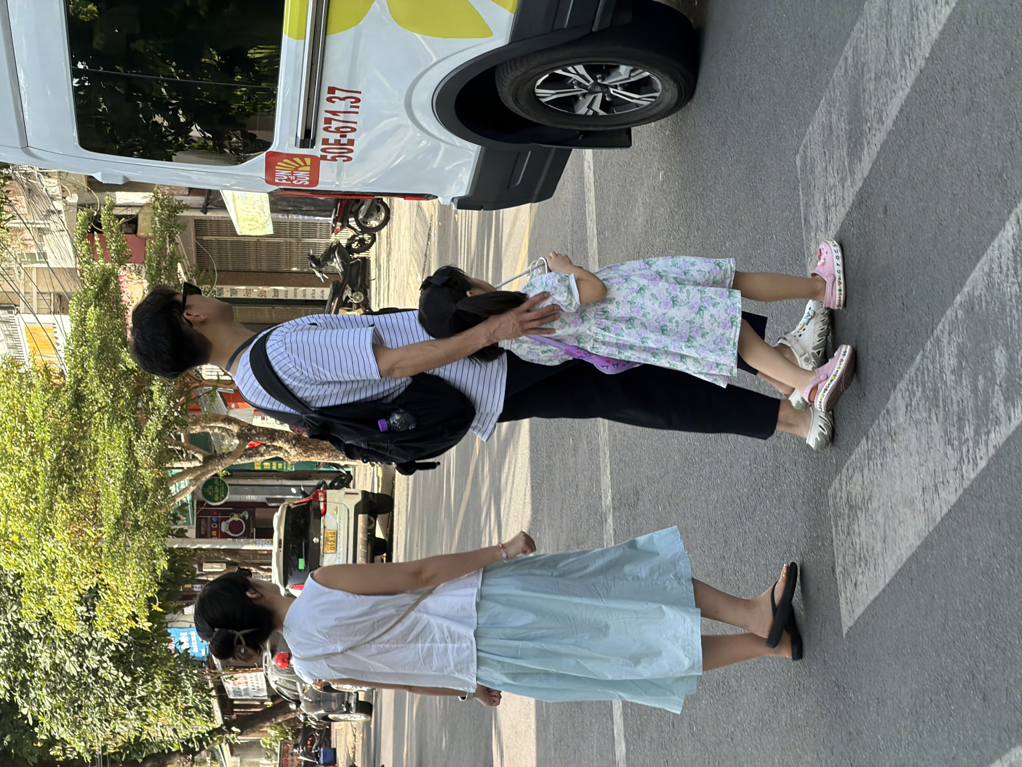In Nha Trang, Vietnam, a family is crossing a street at a crosswalk. A woman in a white sleeveless shirt and a light blue skirt walks ahead, while a man in a striped shirt and black pants walks behind her, holding the hand of a young girl. The girl is wearing a floral dress and pink Crocs. They are near a white van with "FUN ON SUN" branding and the license plate "50E-671.37". Trees line the street, and buildings are visible in the background. The scene is illuminated by sunlight.
