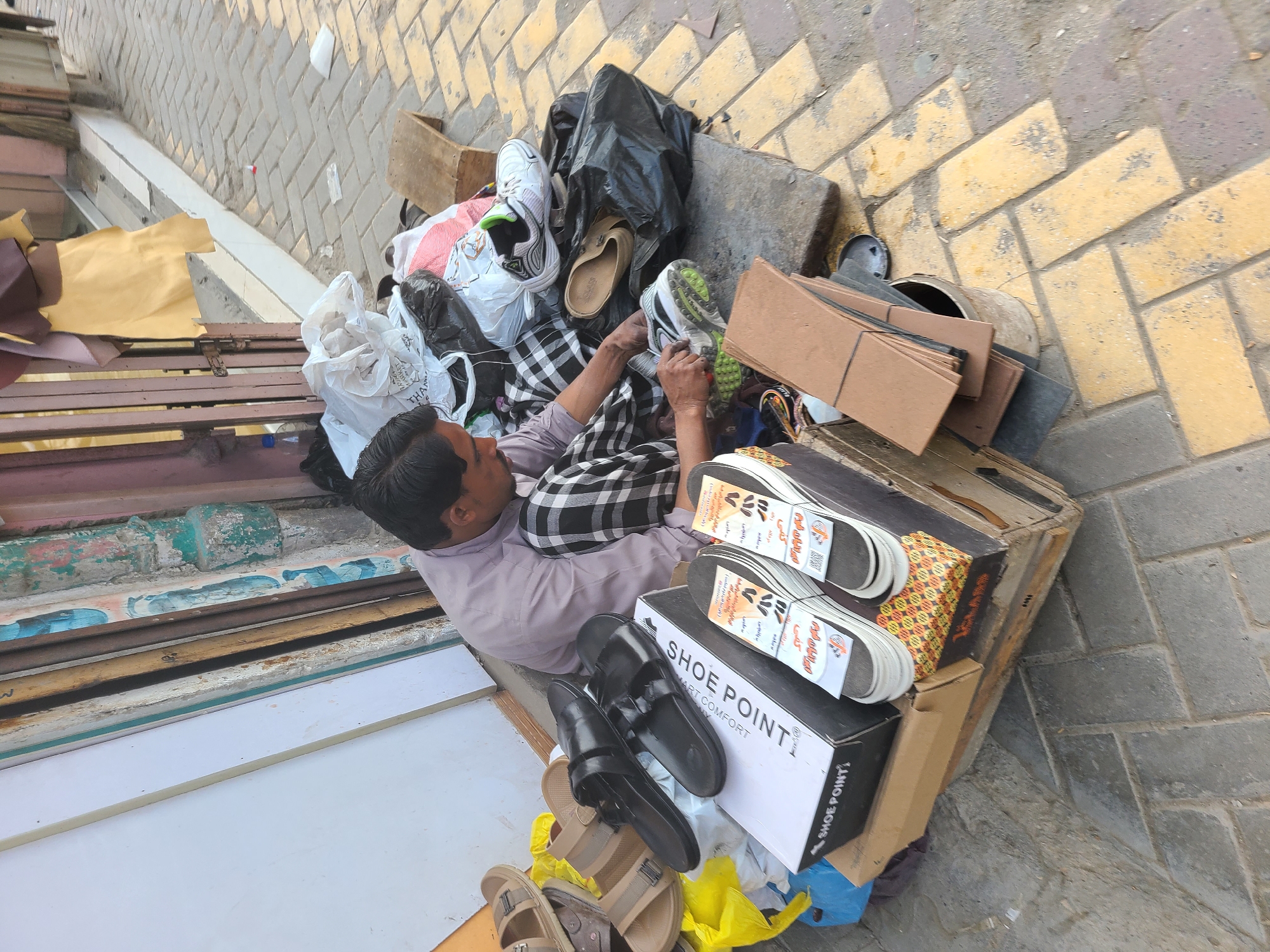 A medium-shot, high-angle photograph shows a man amidst a collection of footwear and various objects, possibly in Quetta City Tehsil, Pakistan.
The man, likely middle-aged with dark hair, is wearing a light purple shirt and a black and white checkered cloth around his lower body. He is handling a white sneaker with green accents.
Around him, there is a jumble of items. These include multiple pairs of shoes and sandals, some stacked on top of "SHOE POINT" boxes, cardboard pieces, plastic bags, and other discarded objects. These items are situated either on the ground or on a low-lying surface alongside a building. The ground appears to be paved with yellow bricks.