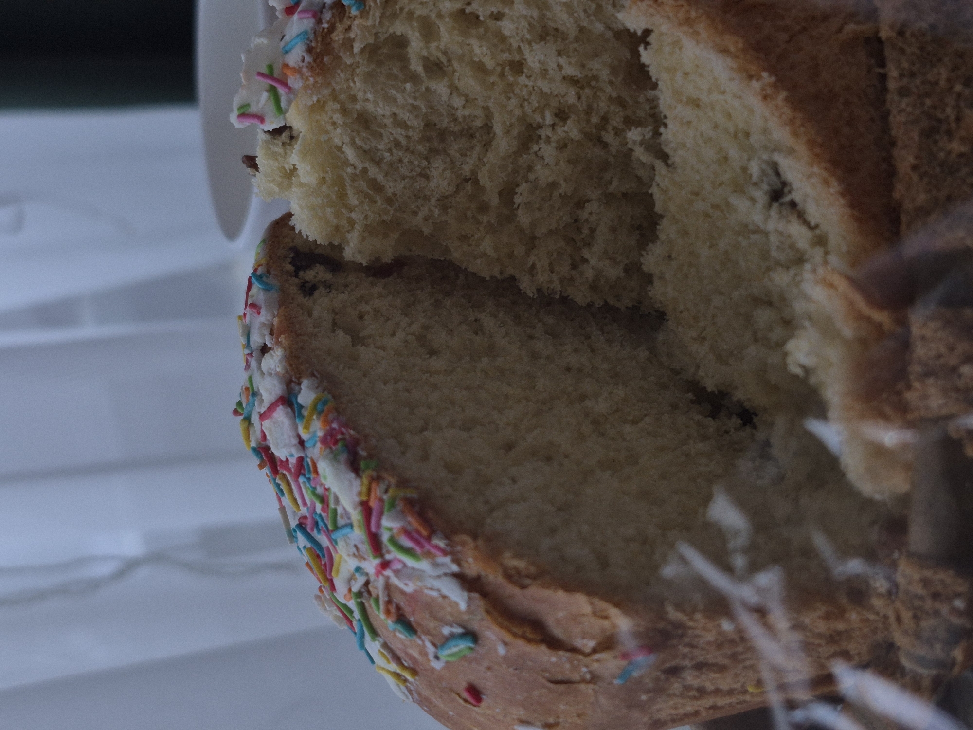 A close-up shot shows a section of kulich, a Russian Easter bread, decorated with white icing and colorful sprinkles. The bread has a dense, spongy texture and appears to be freshly cut. The background is blurry, but hints of a white container and possibly a clear wrapping are visible. The location is likely Chaikovsky, Russia, based on the prompt's instructions.
