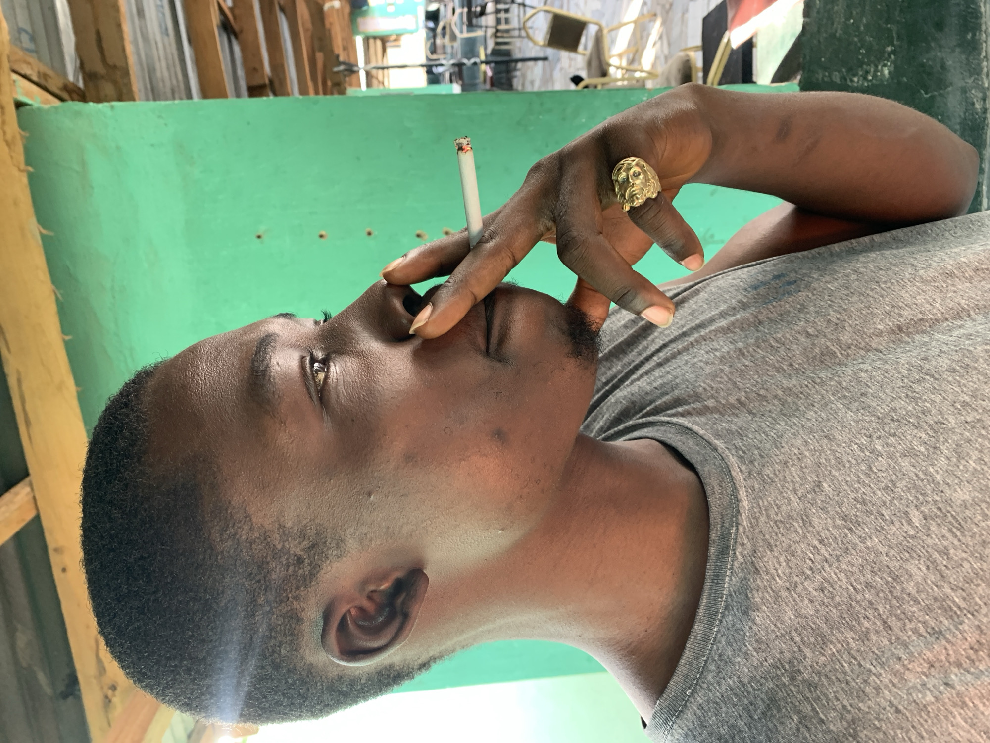 A man is holding a lit cigarette to his mouth. He is wearing a gray t-shirt and a gold ring on his finger. The background is a green wall and some wooden beams. The location is possibly the city of New Stadium, Nigeria, based on user-provided context.