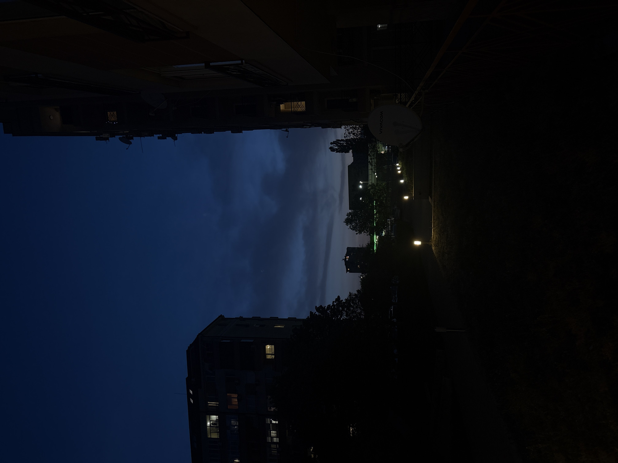 This is an outdoor nighttime shot of buildings and a sky with dark clouds, possibly taken in Sofia, Bulgaria. At the top, an apartment building is visible with a Vivacom satellite dish attached. Down the center, lights illuminate trees and a path, suggesting a park or green space. In the lower portion of the image, another apartment building is visible with a few lit windows. The sky is a deep blue with dark clouds.