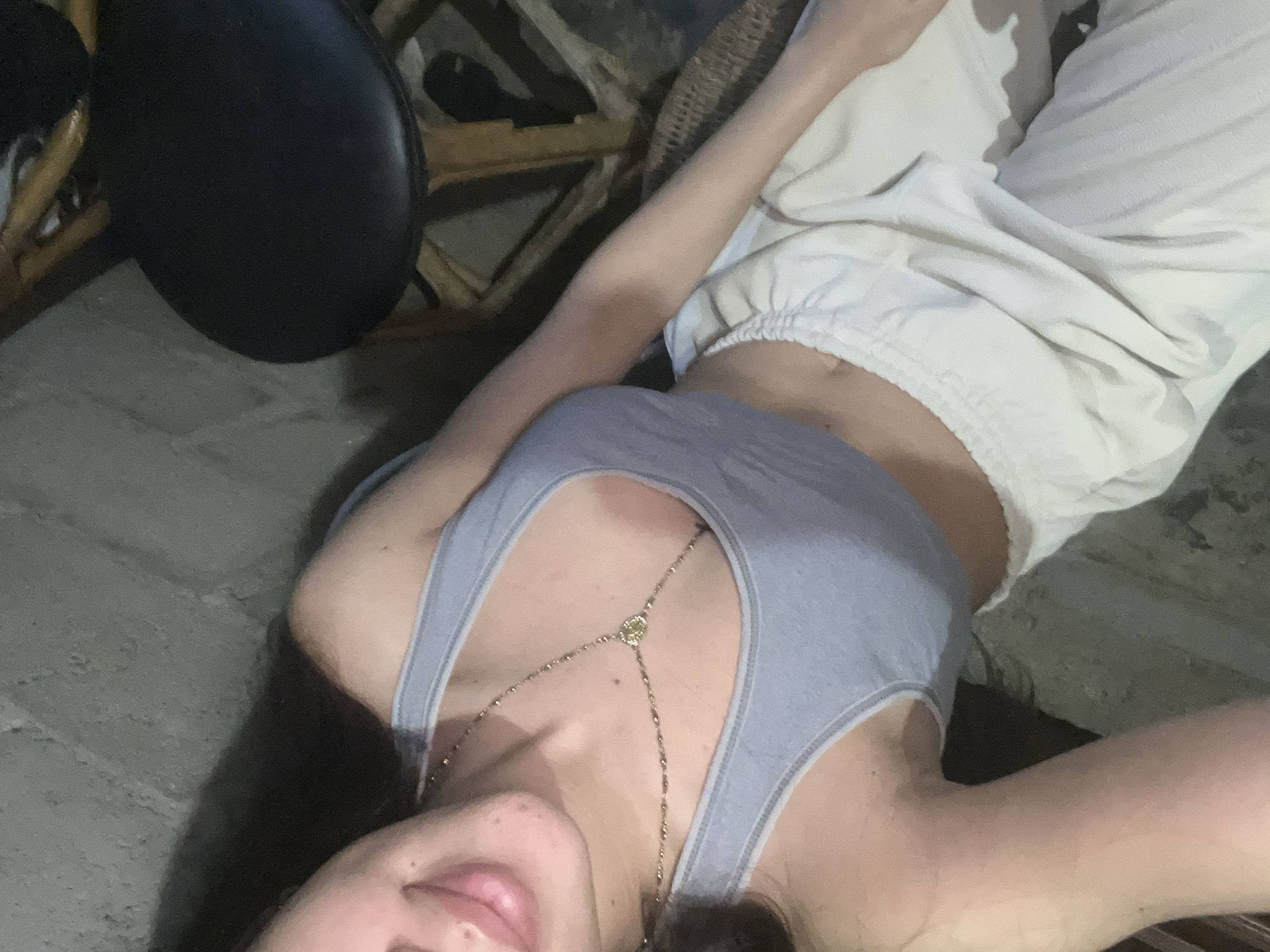 A person, identified as female based on visual cues, is seated. She is wearing a light blue tank top and off-white sweatpants. A delicate gold necklace is worn around her neck, resting on her chest. The background appears to be an indoor setting with textured walls, a stool with a black cushion, and woven furniture. The lighting is dim, casting shadows on the scene. The image is a close-up, focusing on the upper body and lap of the individual.