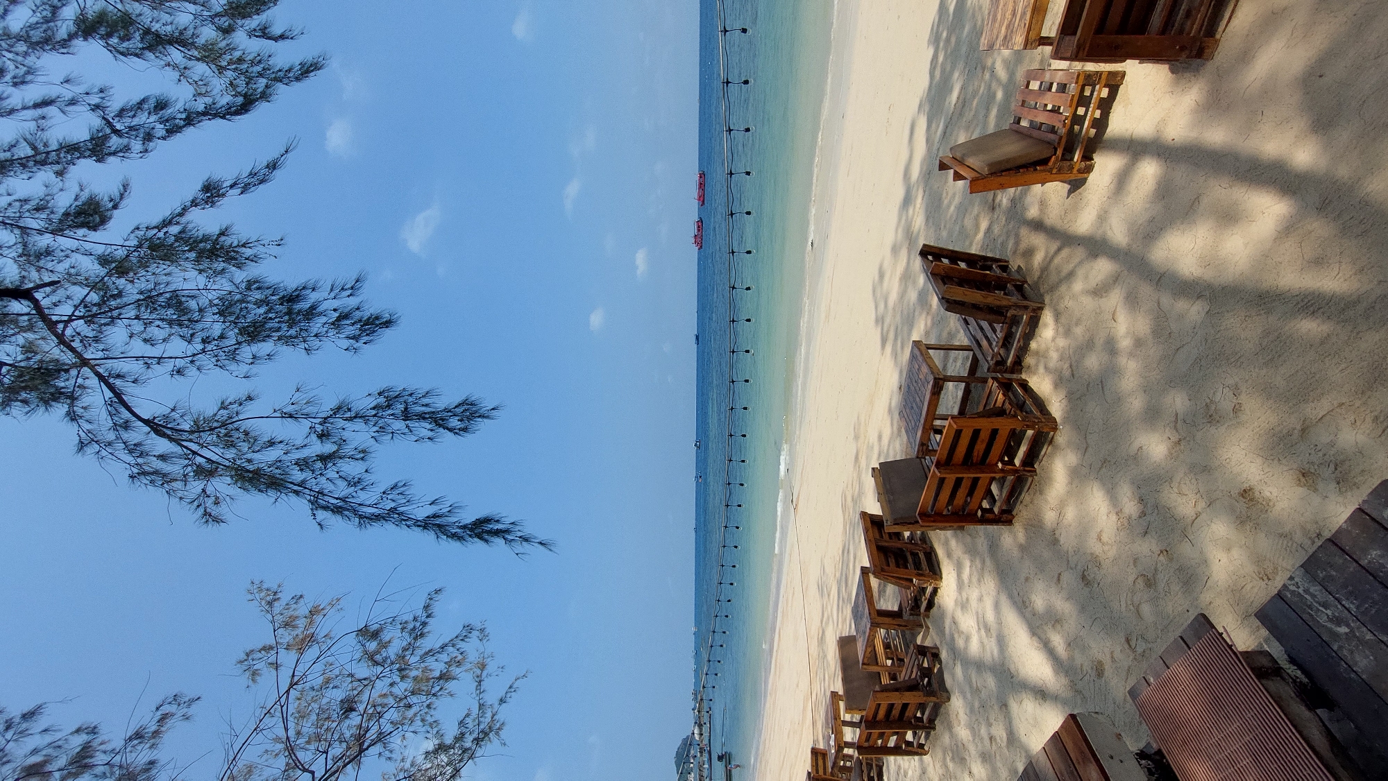 The image shows a beach scene in Koh Tao, Thailand. The view is a long stretch of sandy beach with light wooden lounge chairs and tables arranged in small seating areas along the shore. A line of evenly spaced poles extends out into the clear, light blue-green water, likely marking a swimming or protected area. In the distance, small boats are visible on the water. The sky is a clear, light blue with a few scattered white clouds. Tree branches with sparse foliage frame the left side of the image. Shadows cast by the trees and structures fall across the sand, indicating a sunny day.
