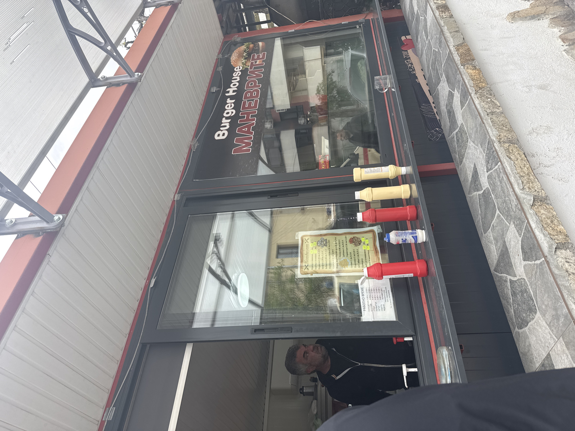 An outdoor food stand, identified as "Burger House МАНЕВРИТЕ" (MANEVRITE) by its black signage featuring a burger graphic, is depicted under a grey corrugated metal awning. The stand has large glass windows, through which a man with dark hair and a beard, wearing a dark top, is visible inside, looking out. The scene is illuminated by daylight, possibly overcast.
On a ledge outside the window, several squeeze bottles of condiments, primarily red (ketchup) and yellow (mustard), are arranged, along with a blue and white bottle, likely salt. A framed menu board, titled "ЦЕНОРАЗПИС" (Price List), is displayed behind the glass, detailing various food items in Cyrillic, including burgers with pork or beef, kebapche, chicken steak, sudzhuk, fried potatoes, pita bread, sauces, and parlenka, with prices listed in Bulgarian lev (лв) and Euro (€).
Another sign specifies the "РАБОТНО ВРЕМЕ" (Working Hours): Monday to Saturday from 8:00 to 17:00, with Sunday being a "ПОЧИВЕН ДЕН" (Day off). The ground next to the stand is paved with grey and tan-colored, irregularly shaped stones. The windows reflect surrounding buildings and the sky. The overall setting suggests a casual, street-side eatery, consistent with its location in Lukovit, Bulgaria.