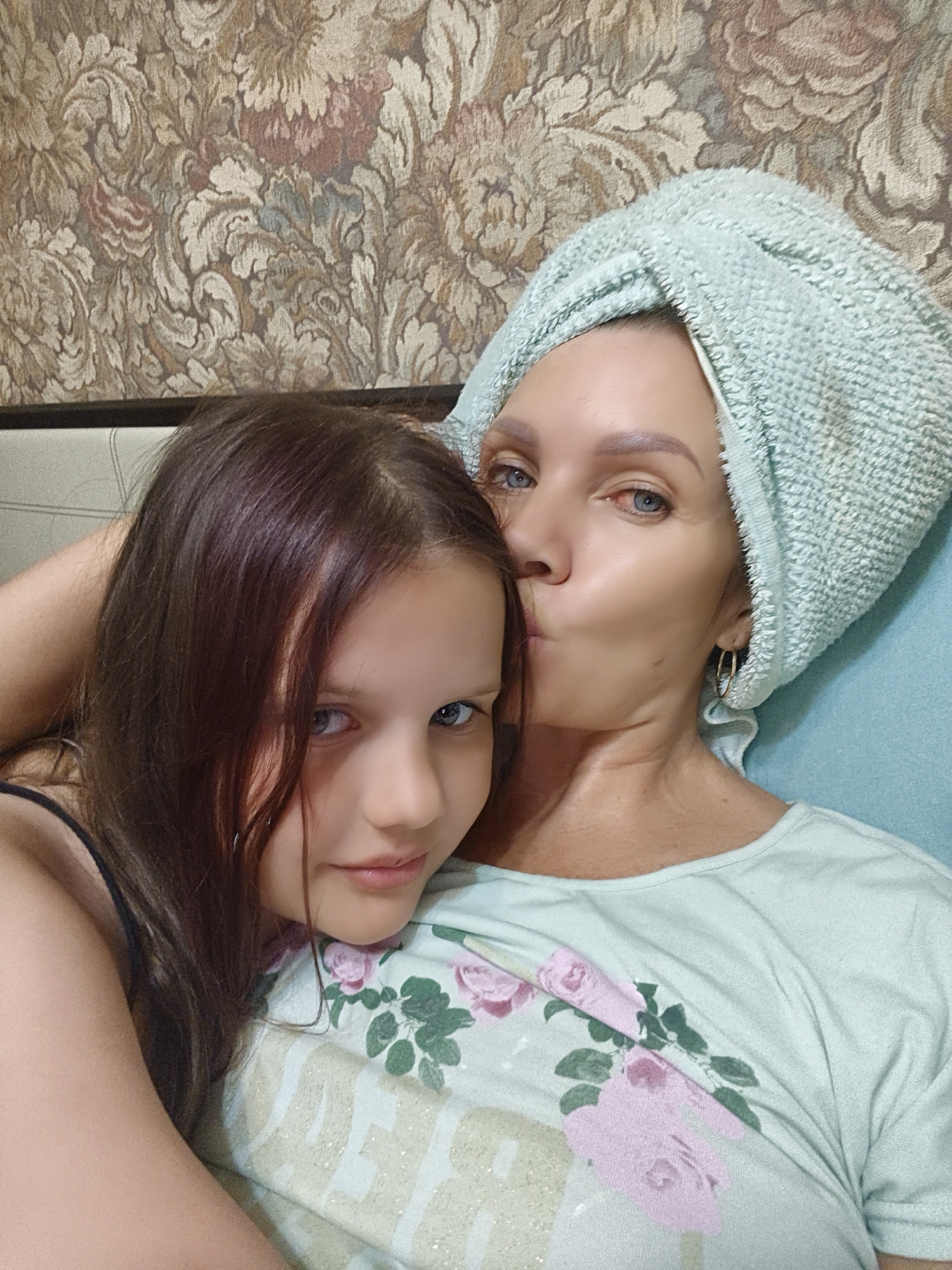 In a close-up, vertical shot, a woman with a light blue towel wrapped around her hair leans in to kiss a young girl. The woman's eyes are closed, and her lips are puckered. She has blue eyes and is wearing a light blue t-shirt with a floral print of pink roses and green leaves. A delicate silver hoop earring is visible in her right ear. The girl has long, dark brown hair that falls over her shoulder and is looking at the camera with a slight smile. Her eyes are also blue, and she appears to be wearing a dark-colored tank top.
They are positioned close together, suggesting a warm and affectionate relationship. The background shows a textured wallpaper with a floral pattern in muted tones of beige, brown, and some hints of pink and gray. The overall lighting is soft, implying it could be daytime or evening indoors. The scene conveys a sense of tenderness and familial love. No text is visible in the image.
