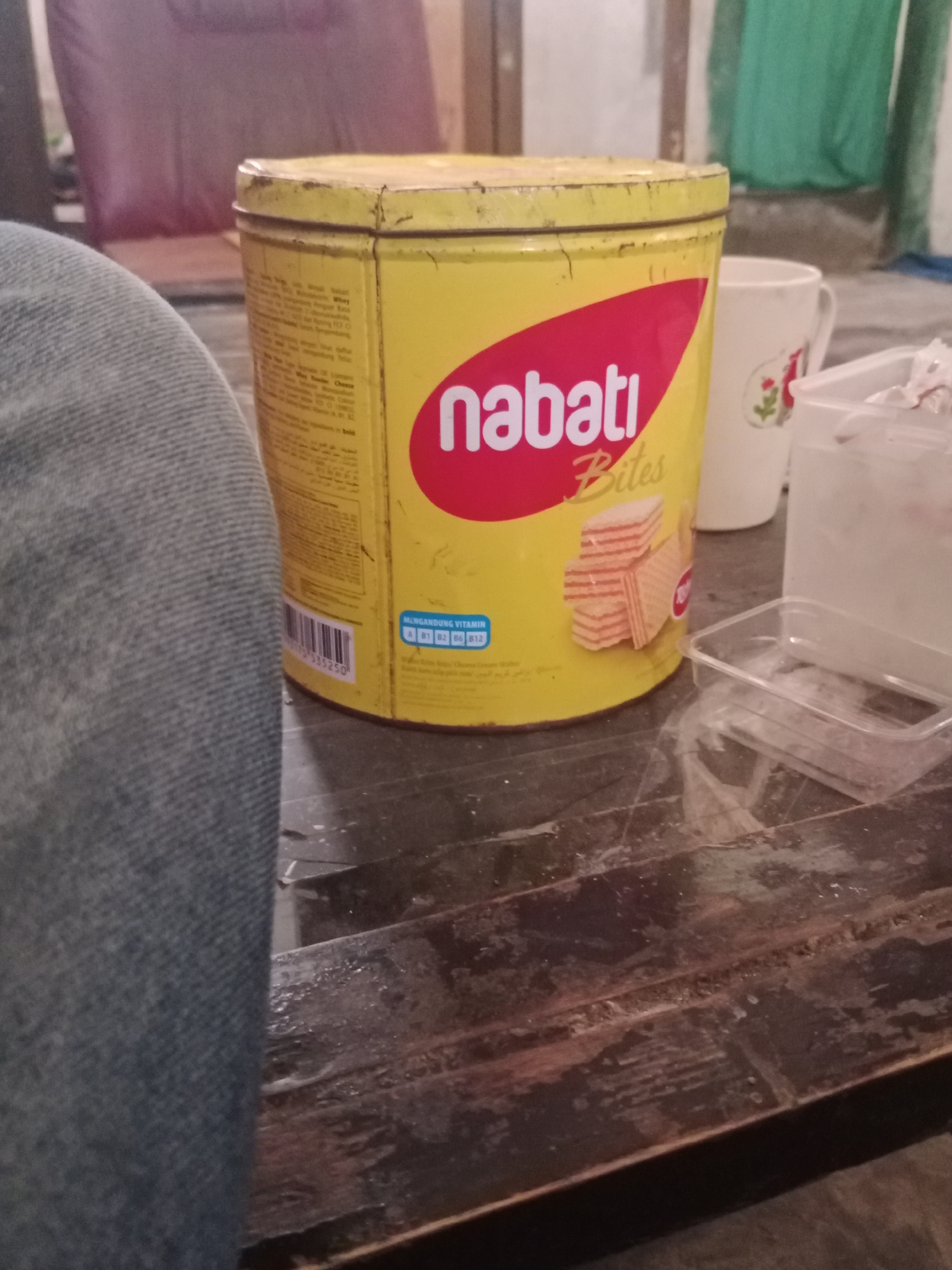 An outdoor, eye-level, medium shot shows a yellow tin of Nabati Bites, a popular wafer snack in Indonesia, positioned on a wooden table. The tin, which is slightly worn, displays the brand's logo and an image of stacked wafers. To the left, a person's leg, clad in blue denim jeans, is visible, partially obscuring the tin. In the background, a white ceramic mug with a floral design and a clear plastic container are also present on the table. The scene appears to be set in a casual, possibly domestic, environment. The background is blurred, but hints of furniture and greenery suggest an outdoor or semi-outdoor location within Ringinrejo, Indonesia.