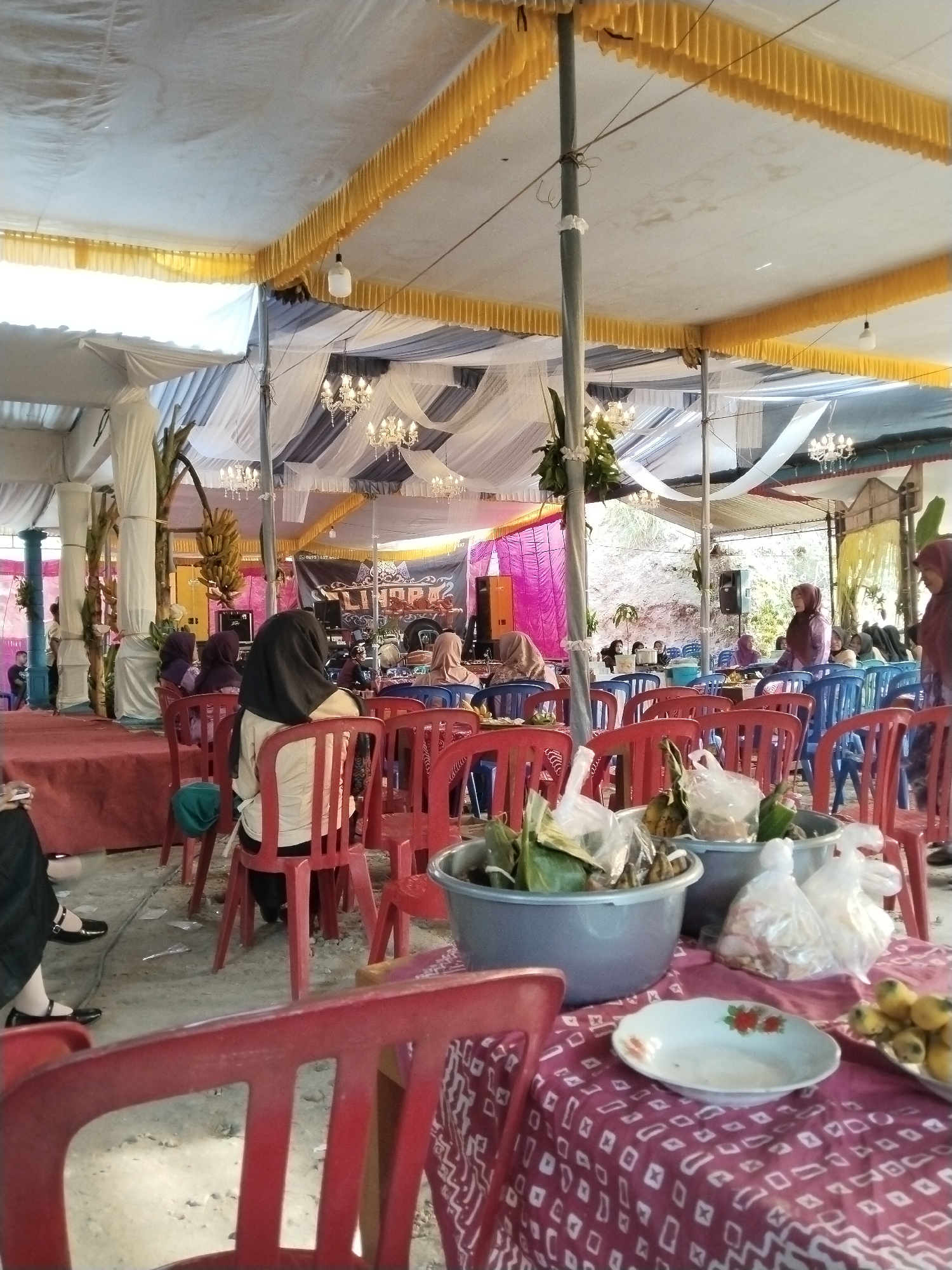 The image depicts an outdoor event space in Jetis, Indonesia, possibly a wedding reception or a similar gathering. A large white tent with yellow trim covers the area, with decorative white fabric draping from the ceiling along with several chandeliers. The ground appears to be a dirt or gravel surface.
In the foreground, several red plastic chairs are visible, with some placed around tables covered with red and white patterned cloths. On one table, a grey basin holds food items wrapped in banana leaves and plastic bags. A white plate with a floral design and a plate of yellow fruit are also present on the table.
A group of people, mostly women wearing hijabs, are seated on chairs, facing a stage area in the background. Some are looking towards the stage, while others are conversing. The stage area is adorned with fabric, banners, and what appear to be musical instruments, suggesting a performance is either in progress or about to begin. In the mid-ground, more rows of blue and red plastic chairs are arranged.
Further back, the natural landscape is visible, with a sloped, rocky hill rising behind the tent. The overall atmosphere appears festive and communal.