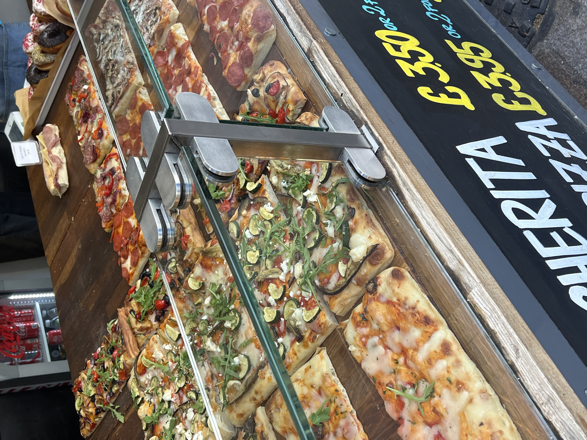 The image shows a display case filled with various types of pizza slices and sandwiches, with a sign that reads "PIZZA MARITA £3.50 or £3.95." The pizza slices have different toppings, including pepperoni, vegetables, and cheese. The display case is made of glass and metal. The scene appears to be at a food vendor in London, United Kingdom, as inferred from the prices in pounds.