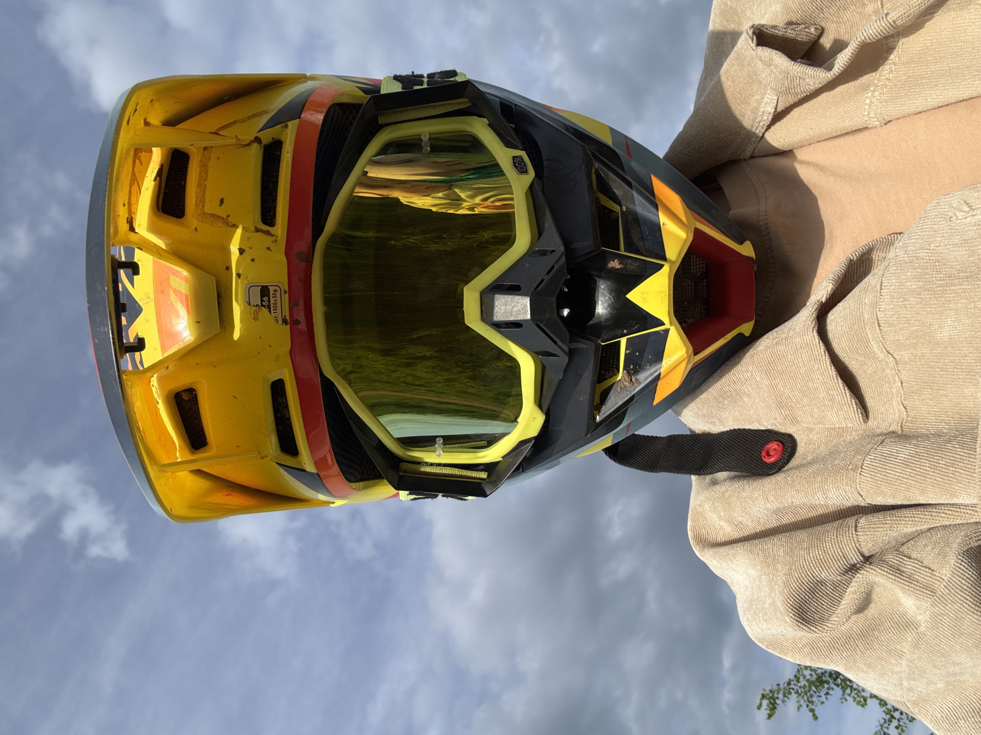 A motocross helmet is positioned in front of a person's chest and shoulder, against a backdrop of sky and clouds. The helmet is predominantly yellow, with black and orange accents, and features attached goggles with a reflective lens. The person is wearing a light brown corduroy jacket. The image orientation is rotated. There is no information available to determine if this image was taken in Votkinsk, Russia.