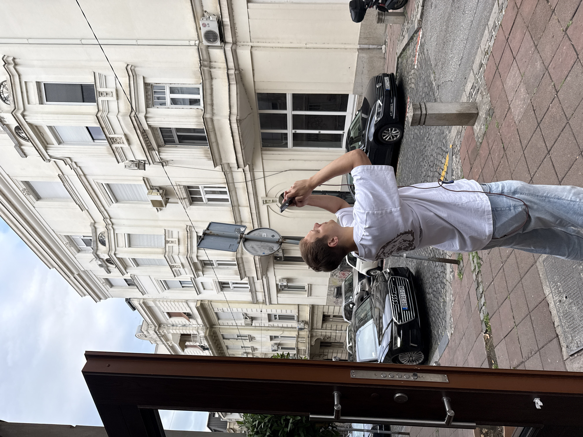 The image depicts a street scene in Belgrade, Serbia. A young person in a white t-shirt and light blue jeans is holding a phone and looking down at it. They are standing on a sidewalk paved with red and grey tiles, next to a street with cobblestone paving. A black car is parked on the street. In the background, there is a multi-story building with cream-colored facade and numerous windows. One air conditioning unit is visible on the building's facade. A street sign with two circular signs is also visible. The scene appears to be during the daytime, under an overcast sky. The overall mood is casual and everyday. The license plate of the black car is visible and reads "BG 3G13 ON".