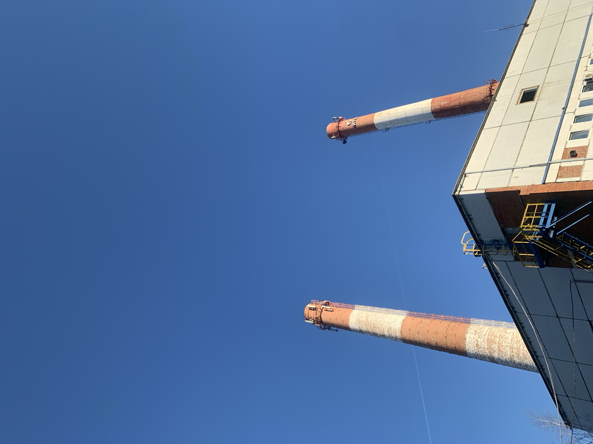 The photograph captures a view looking upwards towards industrial structures against a clear, bright blue sky. Two large chimneys, painted with alternating red and white sections, extend diagonally from the upper right into the center of the frame. A tall, rectangular building with a predominantly white facade is visible on the right side of the image. This structure incorporates brickwork and features windows. Metal platforms and ladders are attached to its exterior, adding to the industrial aesthetic. The chimneys and building appear weathered, with signs of wear and tear visible on their surfaces. A faint contrail can be seen stretching across the sky. The location is Chaikovsky, Russia.