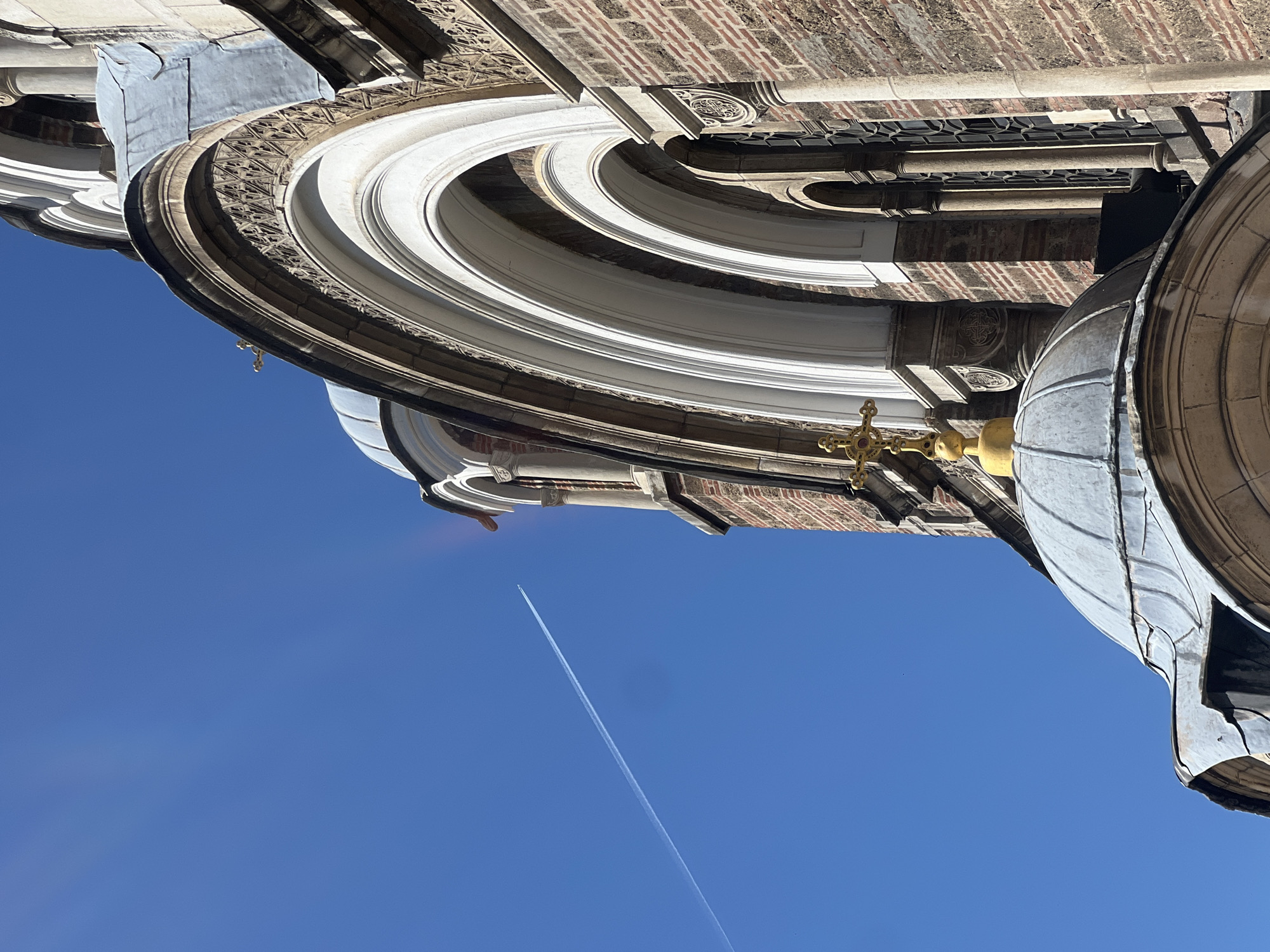 The image shows an upward view of the sky and the architectural details of a building. The sky is a clear blue with a visible contrail from an airplane. Architectural elements include a large dome, several layers of ornate moldings and arches, brickwork, and a golden cross. The building appears to be made of a combination of stone and brick.
The location is the Alexander Nevsky Cathedral in Sofia, Bulgaria.