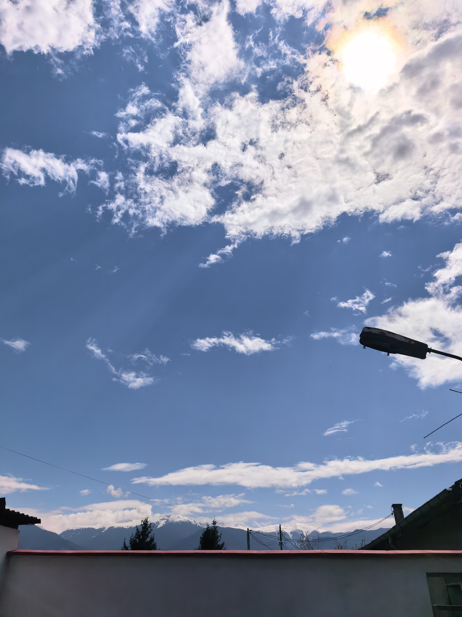 The image depicts a daytime scene in an unknown city location. The sky is mostly blue with scattered white clouds and a visible sun with a faint rainbow halo. A street lamp is positioned in the mid-right foreground. Below, a white wall with a red trim partially obscures a view of distant mountains with snow-capped peaks and coniferous trees. Roofs of nearby buildings are visible behind the wall. Power lines run horizontally across the scene.