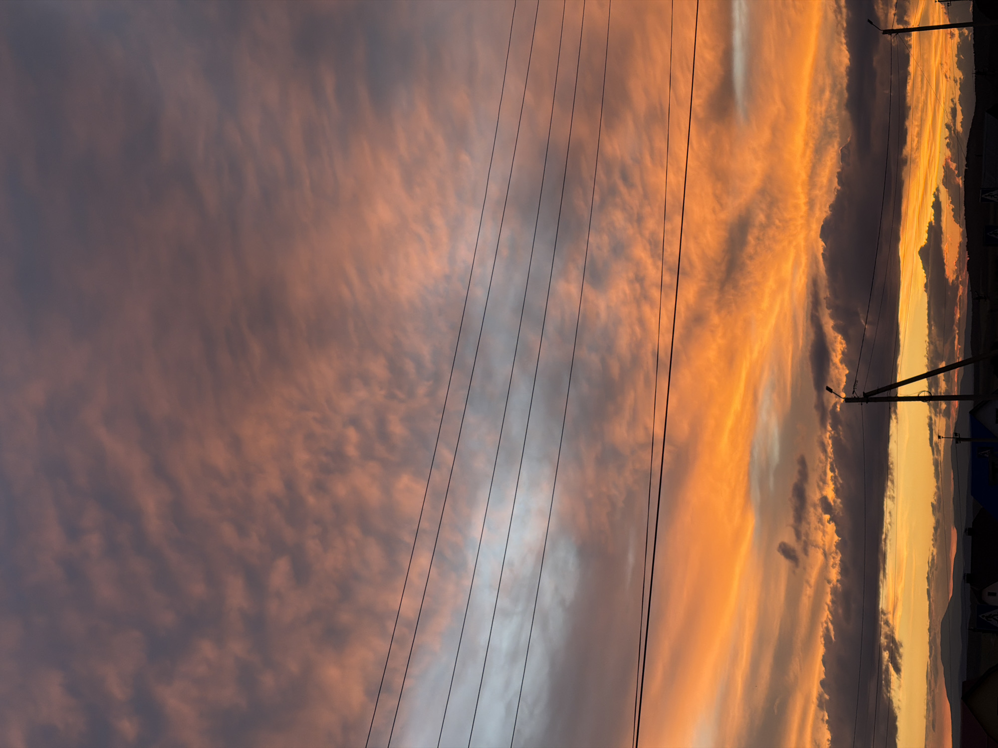 A low-angle, full shot captures a dramatic sunset over a rural landscape, city of Unknown, Unknown. The sky is a vibrant spectacle of color, with fiery oranges and deep reds dominating the lower half, transitioning to softer pinks and grays towards the top. Wispy clouds stretch across the sky, catching the last rays of sunlight. Four parallel power lines stretch horizontally across the frame, dividing the sky. In the lower portion of the image, silhouetted against the bright sky, are the rooftops of several houses and the outline of distant hills. A single utility pole with a lamp is visible on the right side of the frame, adding to the rural, somewhat utilitarian feel of the scene. The overall impression is one of natural beauty juxtaposed with man-made infrastructure.