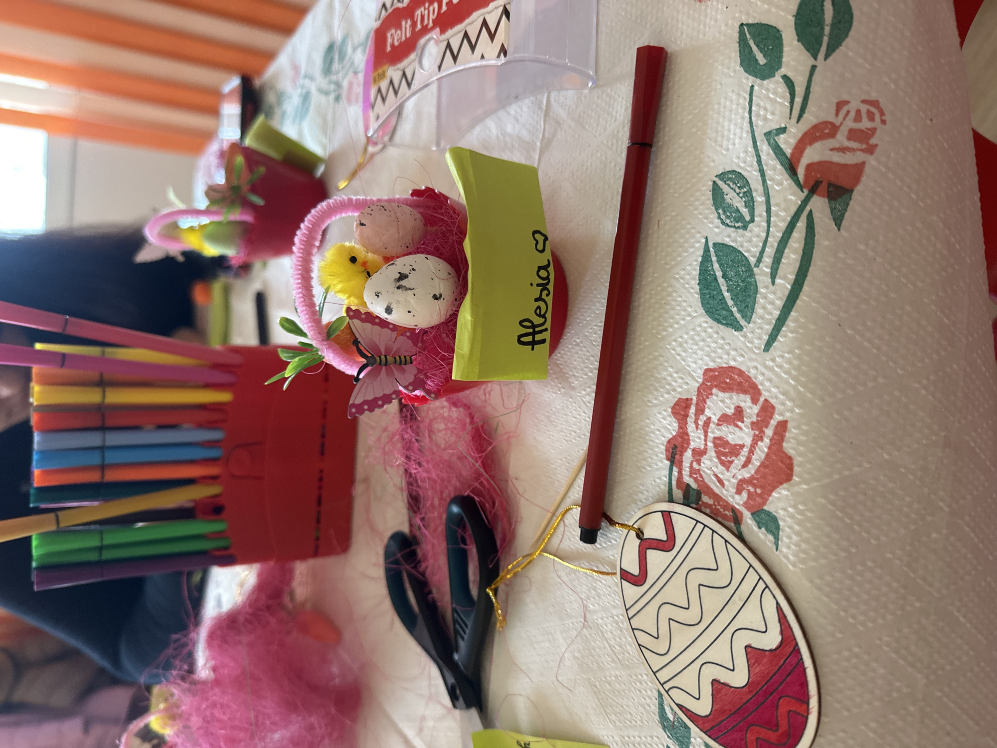 The image shows a tabletop scene with various craft-related objects. There is a container with felt tip pens, small decorative baskets with eggs and chicks, scissors, a marker, and a painted wooden egg ornament. A small name card with "Alessia" is also visible. The tablecloth has a floral pattern with red roses and green leaves. The location is not evident from the image.