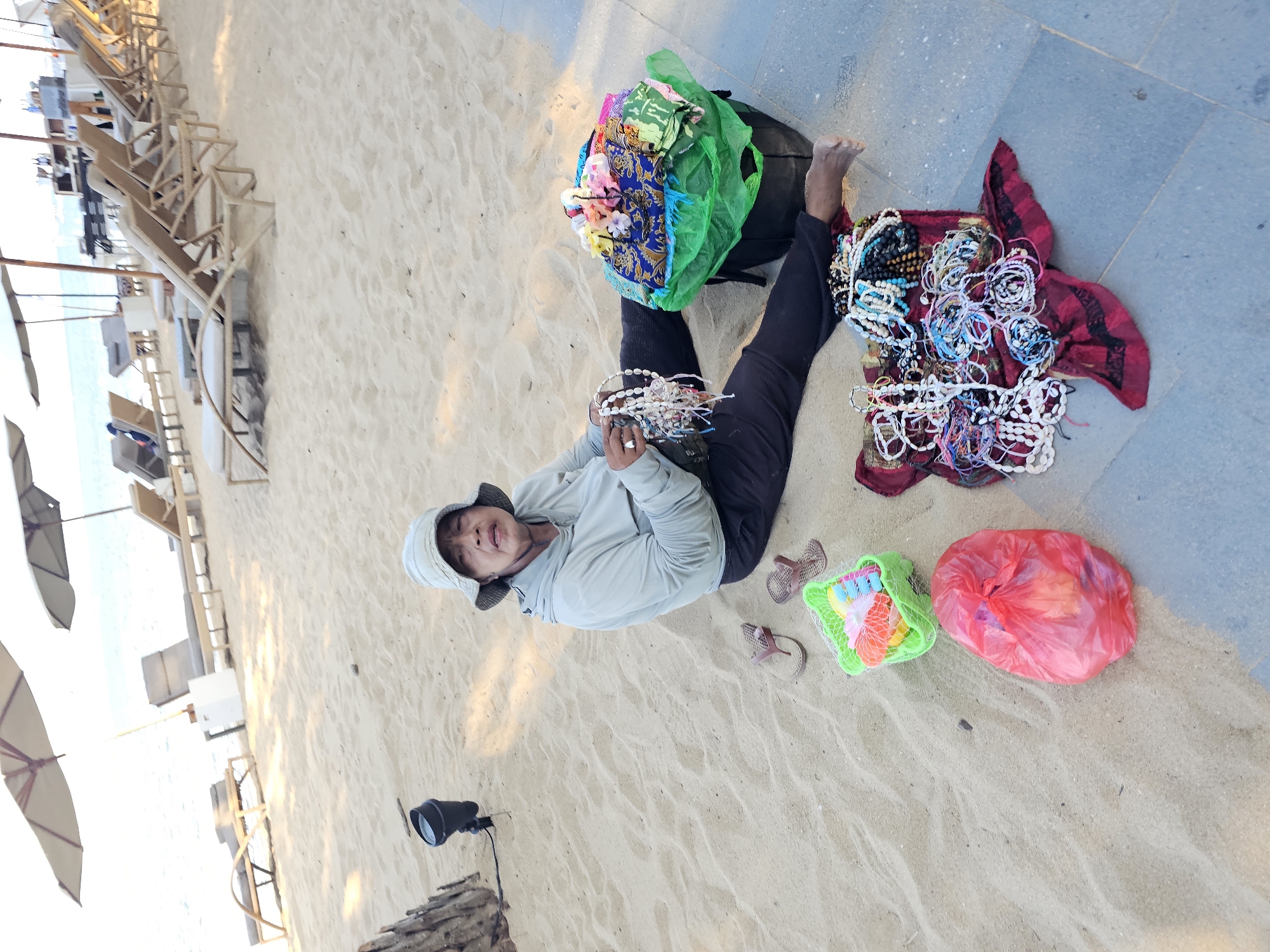 A person sits on a sandy beach, possibly in Denpasar, Indonesia, with several items laid out around them. The person is wearing a light-colored hat and a light-colored hooded sweatshirt with dark pants. They appear to be holding and arranging necklaces or bracelets, with many more such items spread on a red fabric in front of them. To the right of the person is a large green bag filled with colorful fabrics and small flower decorations. In the sand to the left of the person are a pair of flip-flops, a green plastic basket containing colorful plastic toys, and a red plastic bag. In the background, lounge chairs and umbrellas are set up on the beach, with the ocean visible in the distance. A black light fixture is visible on the sand to the left of the person.