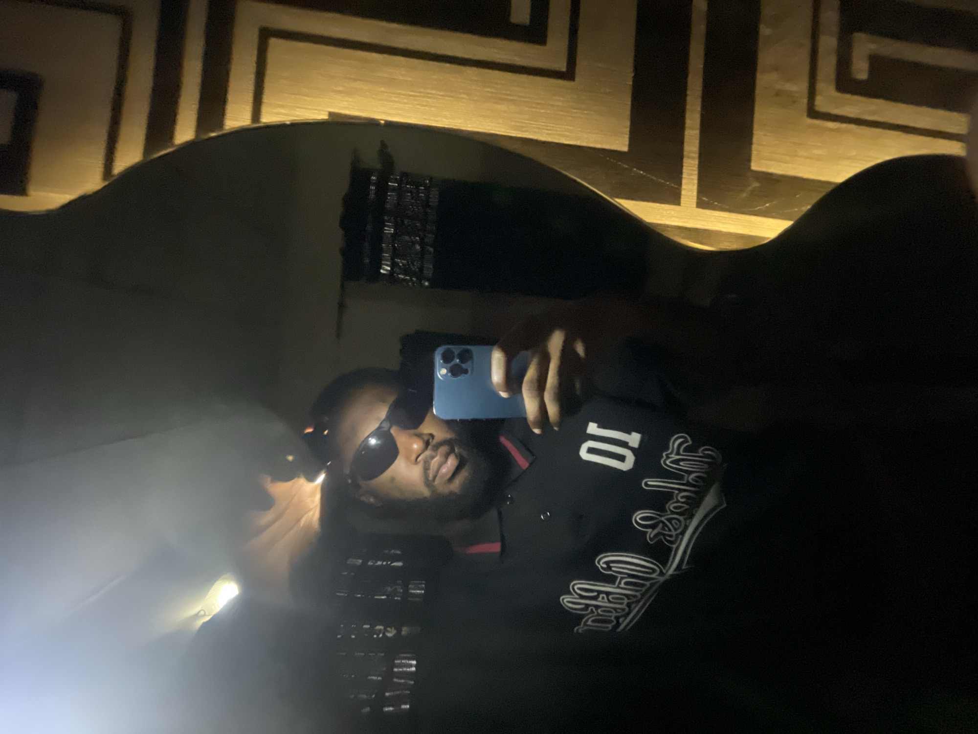 A man is taking a selfie in a mirror. He is wearing a black polo shirt with a white graphic design on the front and sunglasses. He is holding a blue smartphone in his right hand. The background appears to be a room with a decorative wall and a light fixture. The image is dark and grainy, suggesting it was taken in low light conditions.