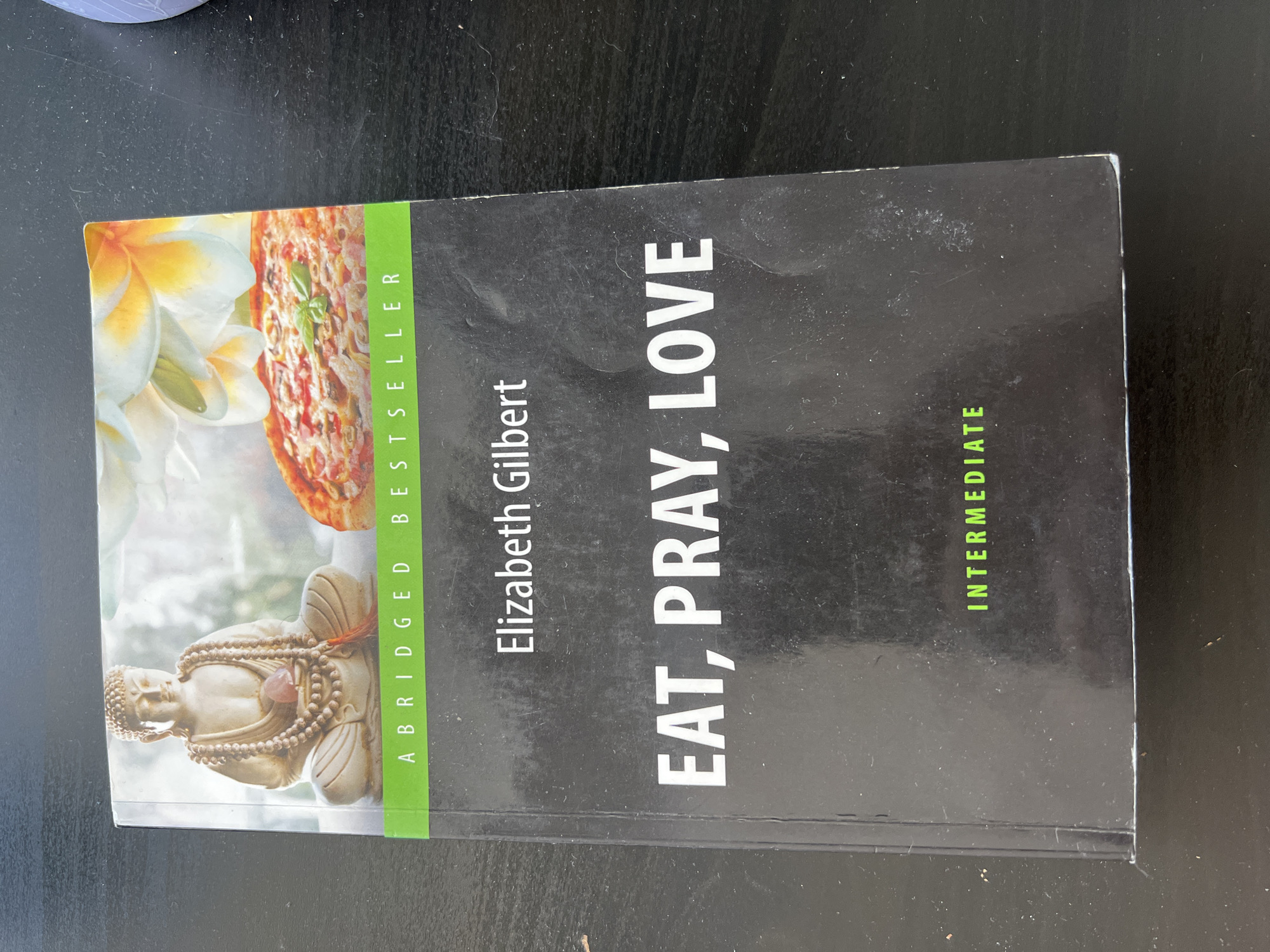The image shows the cover of the book "Eat, Pray, Love" by Elizabeth Gilbert. The cover features a split image: the left side depicts a serene Buddha statue with a mala and a pink stone, while the right side shows a pizza with basil and flowers in the background. A green band across the top of the cover reads "ABRIDGED BESTSELLER". The author's name, "Elizabeth Gilbert", is printed in white below the top image, followed by the title "EAT, PRAY, LOVE" in large white letters. At the bottom of the cover, the word "INTERMEDIATE" is written in green. The book is lying on a dark, textured surface, likely a table. There is no indication of the city of Novosibirsk, Russia, or any other specific location.