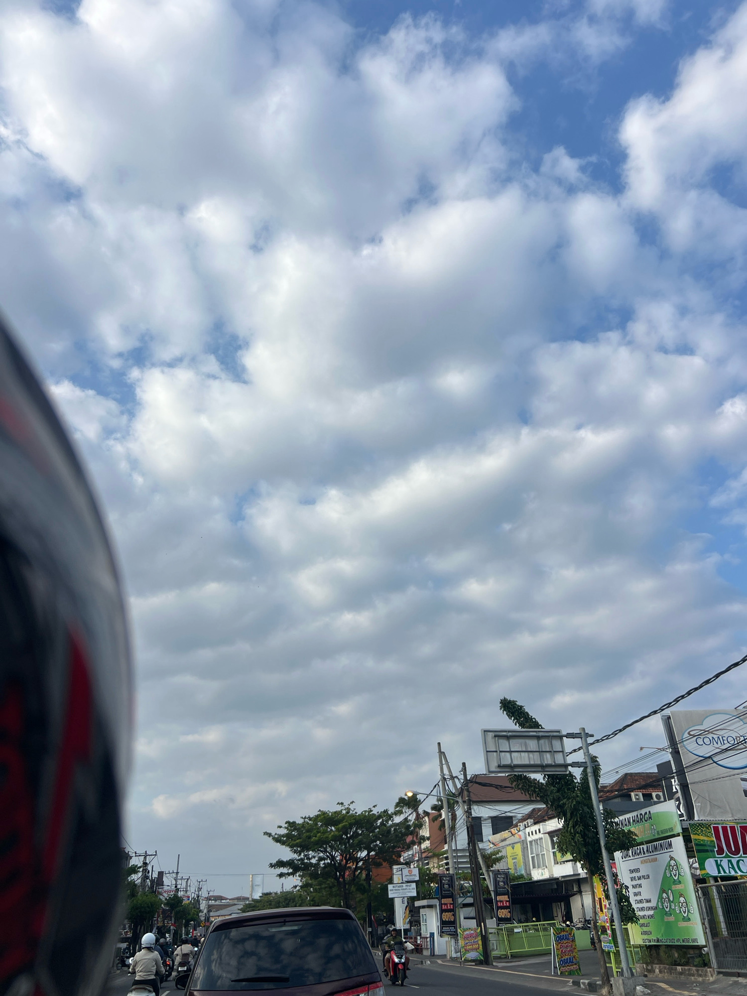 In Denpasar, Indonesia, a street scene unfolds under a partially cloudy sky. The foreground is dominated by the blurred, close-up view of a motorcycle helmet, suggesting the photo was taken by a rider. The helmet is dark with red and white accents. Beyond the helmet, the street is bustling with traffic. Several motorcycles are visible, carrying riders wearing helmets. A dark-colored car is in the lane ahead, reflecting the sky and surrounding buildings.
On the sides of the street, shops and buildings are lined up. One shop displays a prominent green sign advertising "JUAL KACA & ALUMINIUM" (Selling Glass & Aluminum) with illustrations of cartoon figures. Other signs, some featuring percentages like "60%", are also visible, indicating commercial activity. Utility poles with a network of wires crisscross the sky above the street. Trees with green foliage dot the landscape, adding to the urban environment. The overall impression is of a typical day in a busy Indonesian city, with traffic, commerce, and everyday life in motion.