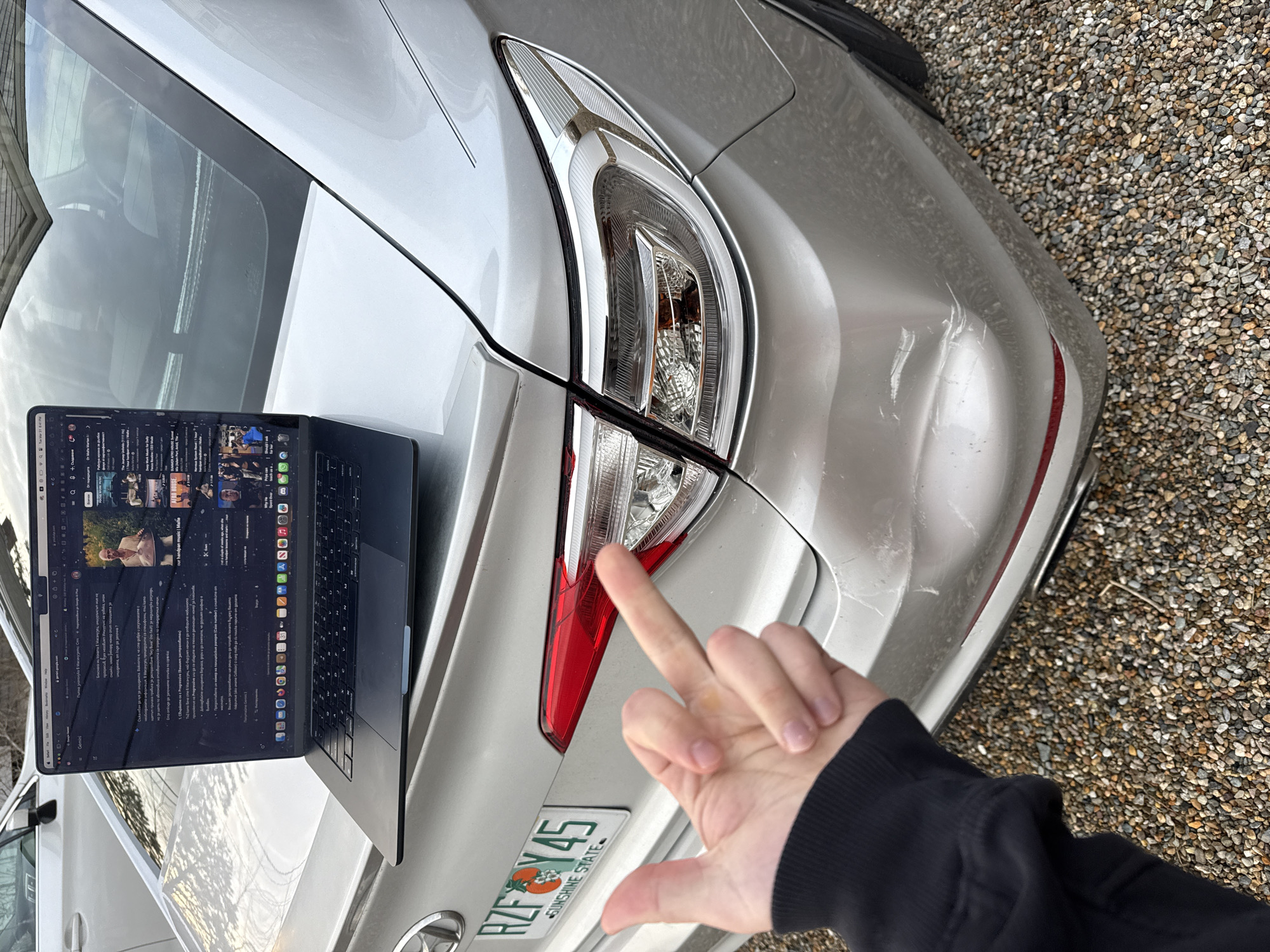 A detailed, close-up, rotated shot shows the rear end of a silver car parked on a gravel surface. The car's right taillight appears cracked. A hand with fair skin, wearing a black sleeve, points towards the taillight. A laptop is positioned on the car's trunk, displaying a webpage with a mixture of text and video thumbnails. A license plate is visible on the lower portion of the car, reading "RZF Y45" and "SUNSHINE STATE". The location is unknown, but the license plate suggests Florida, United States of America.