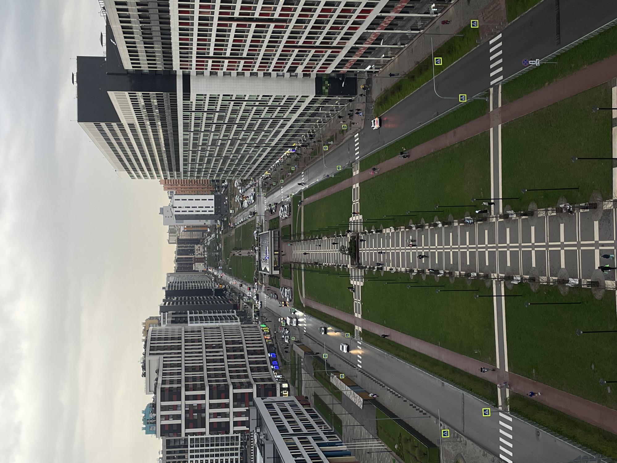 The image depicts a long, wide boulevard flanked by tall buildings in an urban area, potentially in an unknown city in Russia. The boulevard features a central green space with parallel pedestrian walkways adorned with symmetrical lamp posts and seating areas. On either side of the green space are roadways with vehicles and pedestrian crossings. High-rise buildings with repetitive grid-like architectural designs line the streets. The sky is overcast, suggesting a cloudy day. The scene is captured from an elevated perspective, providing an overview of the city landscape.