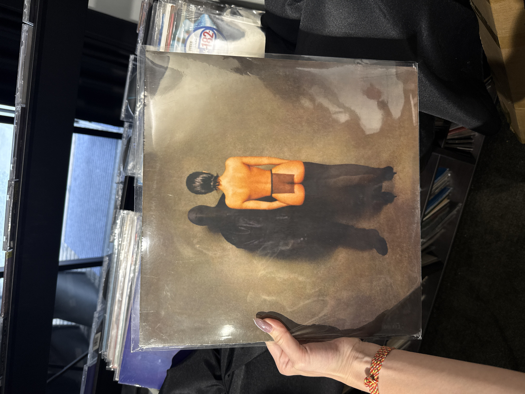 A vinyl record album in a protective plastic sleeve is held in front of a person wearing a black shirt and a multi-colored bracelet on their right wrist. The album cover features a stylized image of a person lying down, viewed from the back, with a rectangular area covering their buttocks. The person's head is visible, along with a dark shadow extending from their body. The background is a muted brown color. To the left, a shelf unit is filled with CD cases, and a window is visible through the shelving. To the right, a black fabric and a cardboard box are present. The scene appears to be indoors, possibly in a music or record store. The Jing'an District, China, is not discernible from the image.