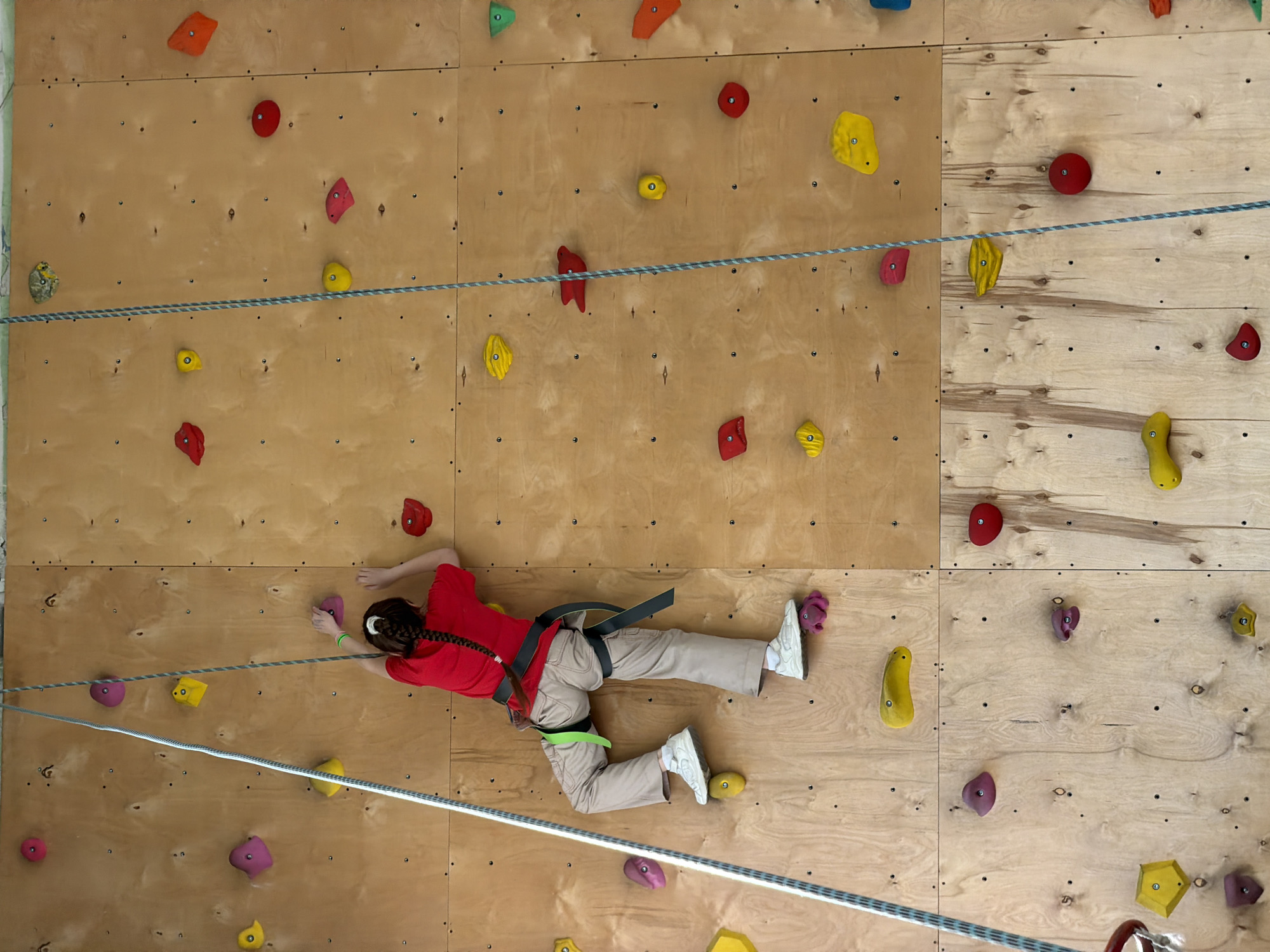A young person is climbing an indoor climbing wall. The wall is made of wood panels and has various colorful climbing holds attached. The person is wearing a red shirt, tan pants, and white shoes, and they are secured with a harness and rope. The rope runs horizontally across the wall. The climbing wall is located indoors, likely in a gym or recreational facility. There is no readily available information to confirm if this image was taken in Izhevsk, Russia.