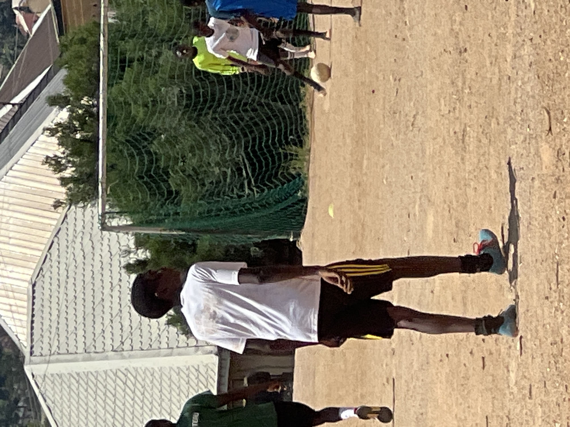 In Jos, Nigeria, a group of young males is engaged in a game of soccer on a dirt field. A tall male in a white t-shirt and black shorts with yellow stripes is the primary subject, facing away from the camera and appearing to be mid-kick as he interacts with the soccer ball. To his left, another male in a green t-shirt and black shorts is partially visible. In the background, a soccer net is set up, with several other individuals in athletic attire present. The environment features a building with a textured, grey roof in the background, and a sparse scattering of green vegetation. The lighting suggests it is daytime, with sunlight casting shadows on the dusty ground.